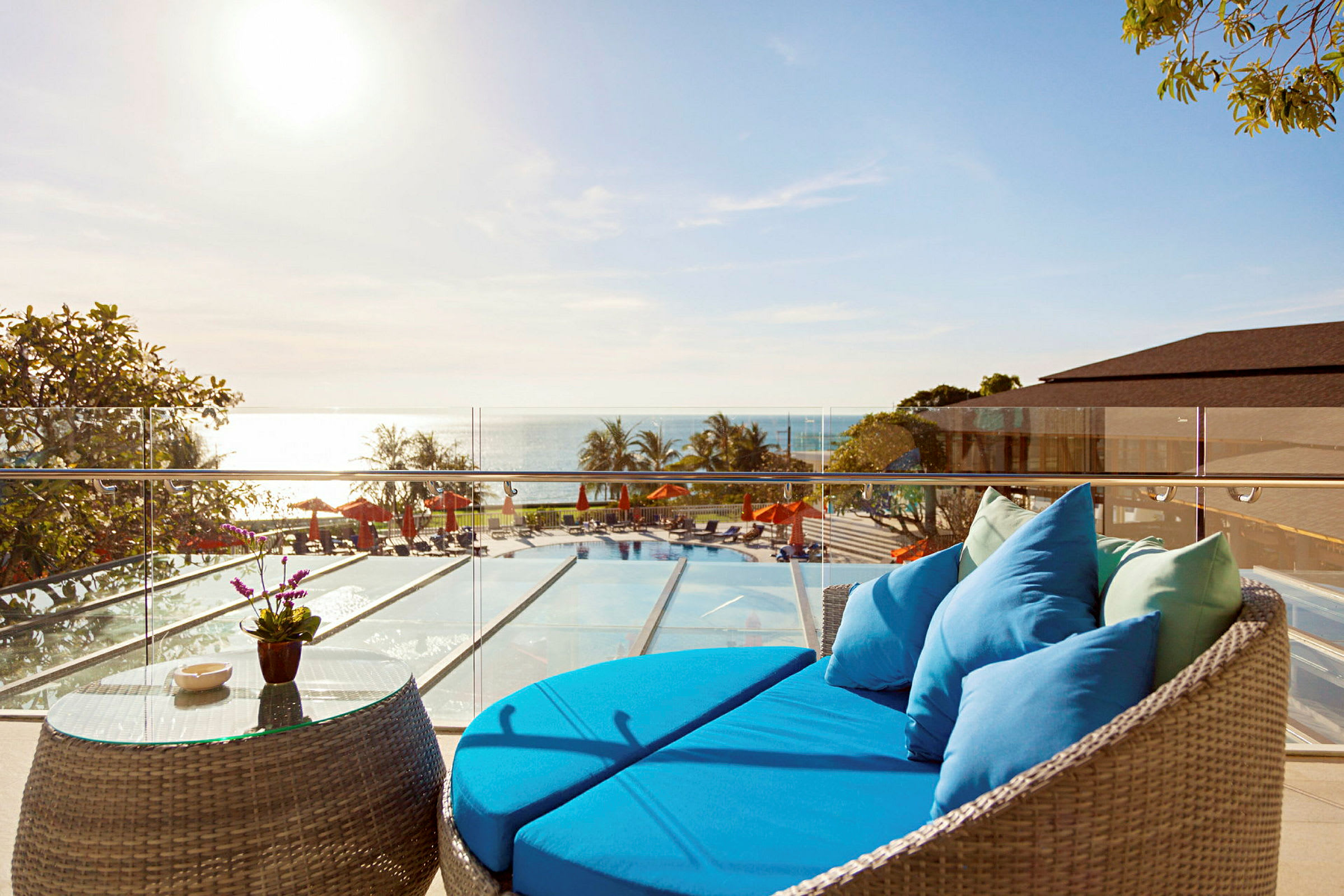 En terrass med en blå soffa och ett glasbord, med utsikt över en pool, havet och solstolar under parasoll. (AI-genererad text)