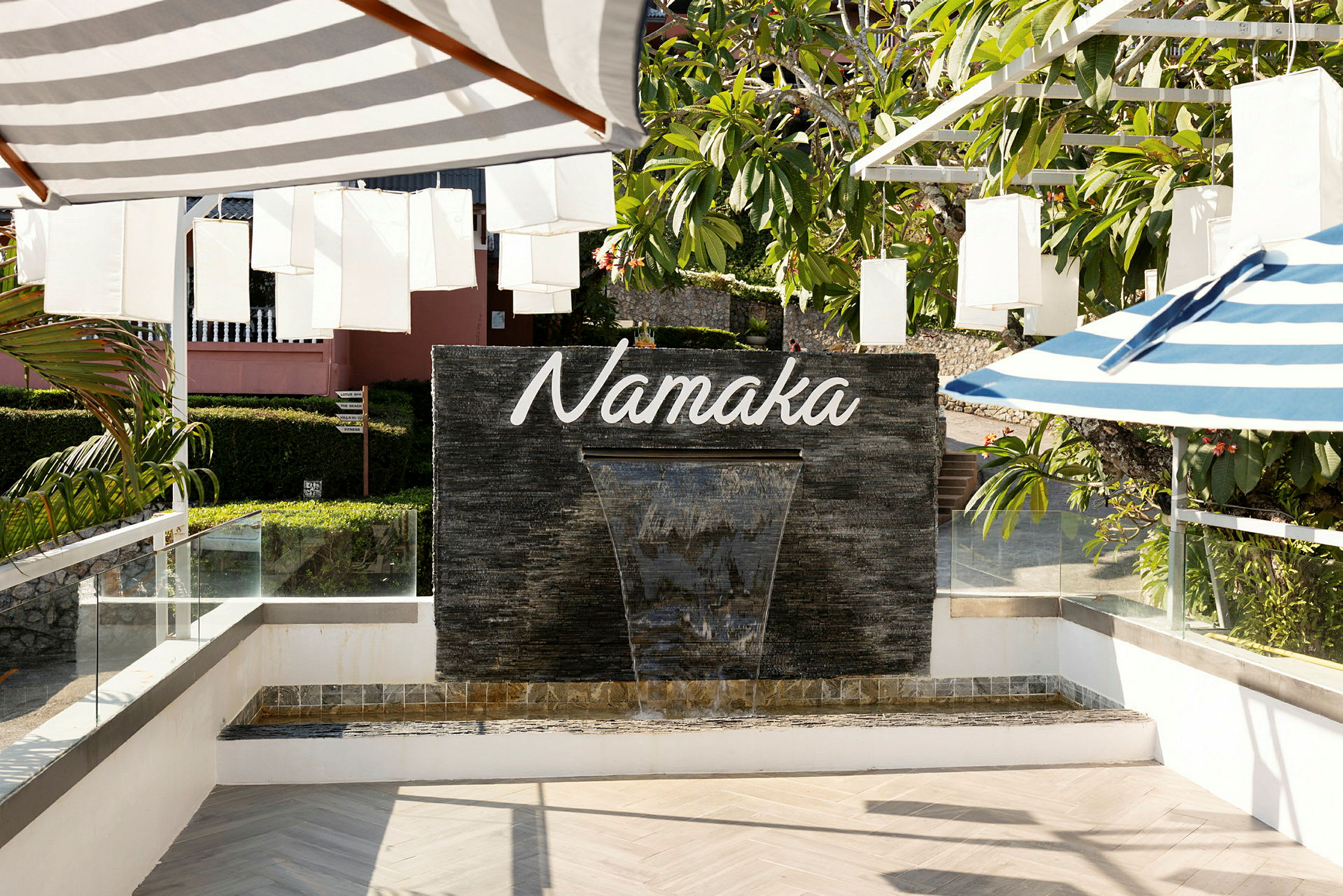 Fontän med texten "Namaka" på en svart stenmur, omgiven av grönska och skuggande paraplyer. (AI-genererad text)