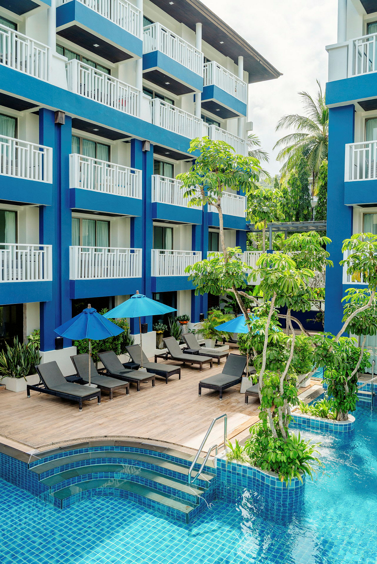 Ett hotell med blå fasad, pool, solstolar och parasoller omgiven av grönska. (AI-genererad text)