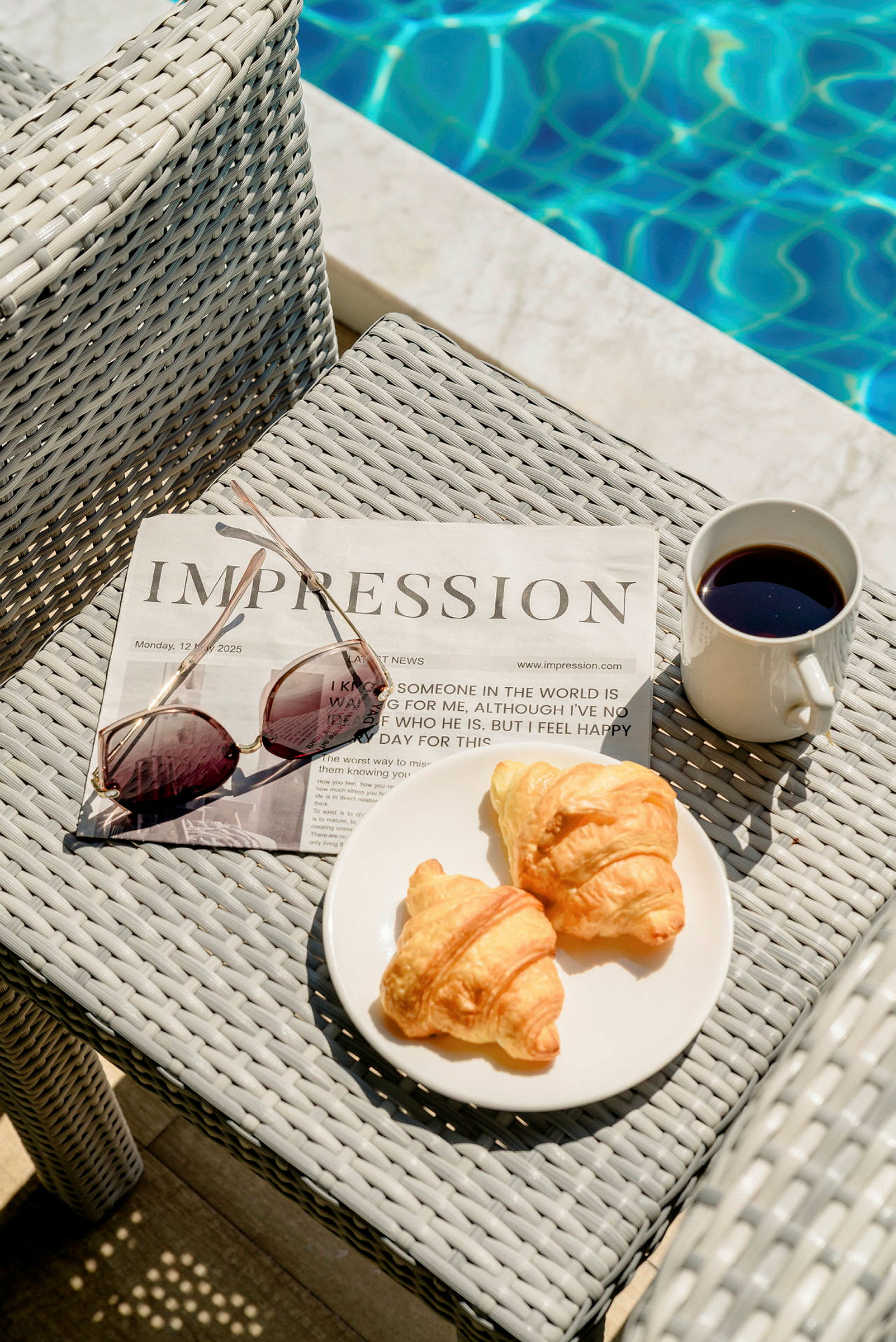 En tidning och solglasögon på en bordsyta bredvid en tallrik med två croissanter och en kopp kaffe, med en pool i bakgrunden. (AI-genererad text)