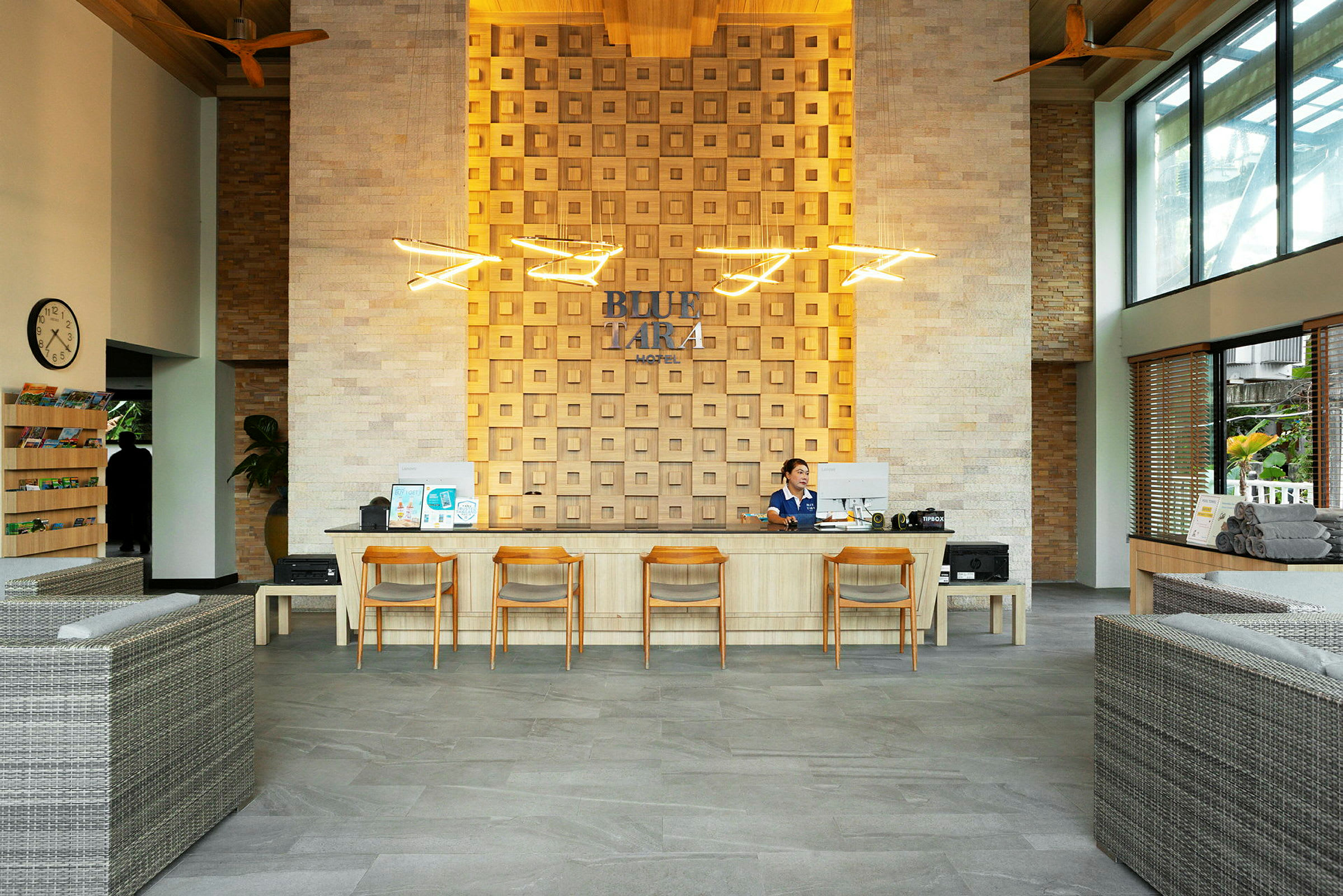 En reception med träbord och fyra stolar. En person arbetar vid receptionsdisken. Väggarna är dekorerade med kvadratiska paneler och modern belysning. (AI-genererad text)