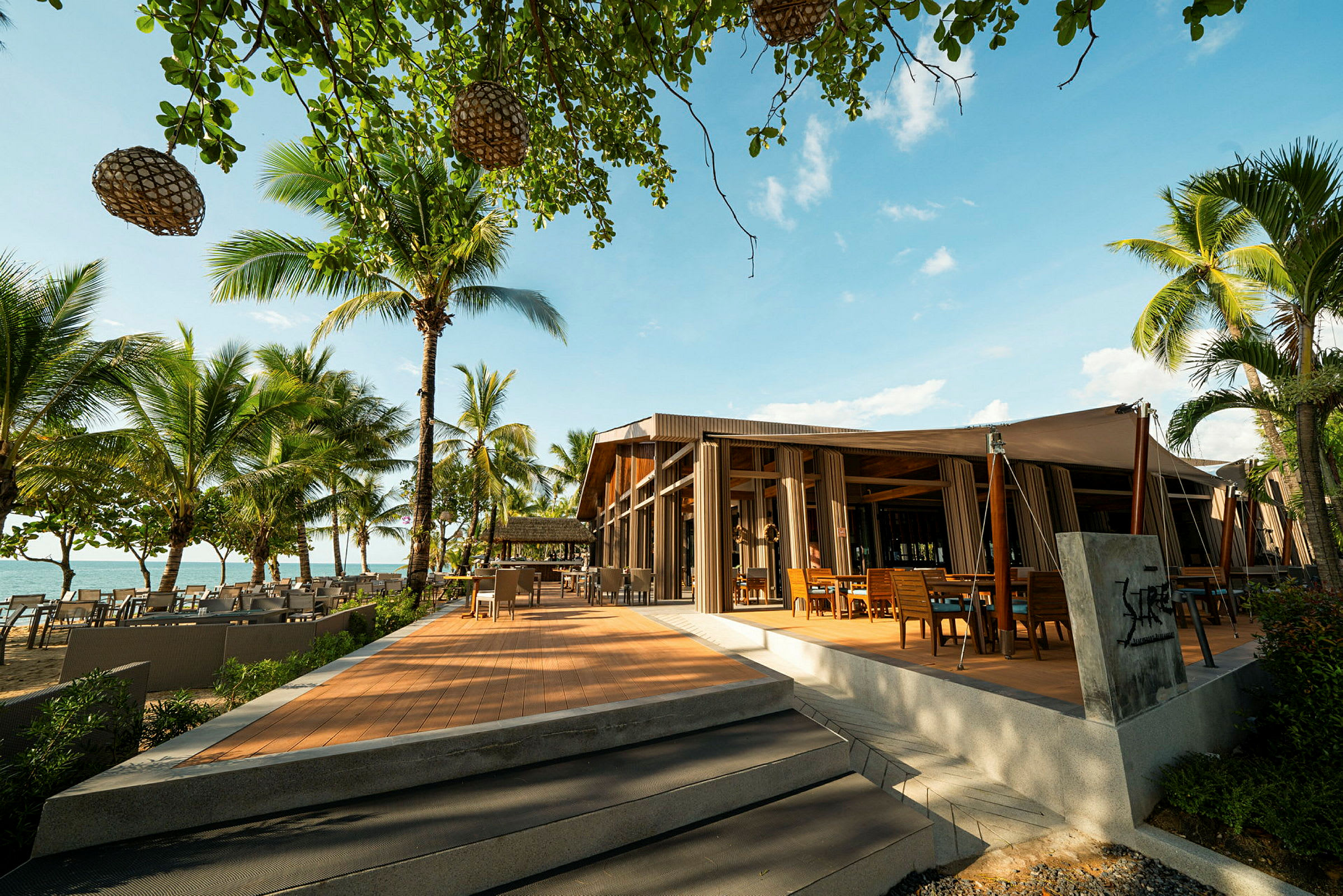 En trevlig restaurang med trädetaljer syns bredvid stranden, omgiven av palmer och en klar blå himmel. (AI-genererad text)