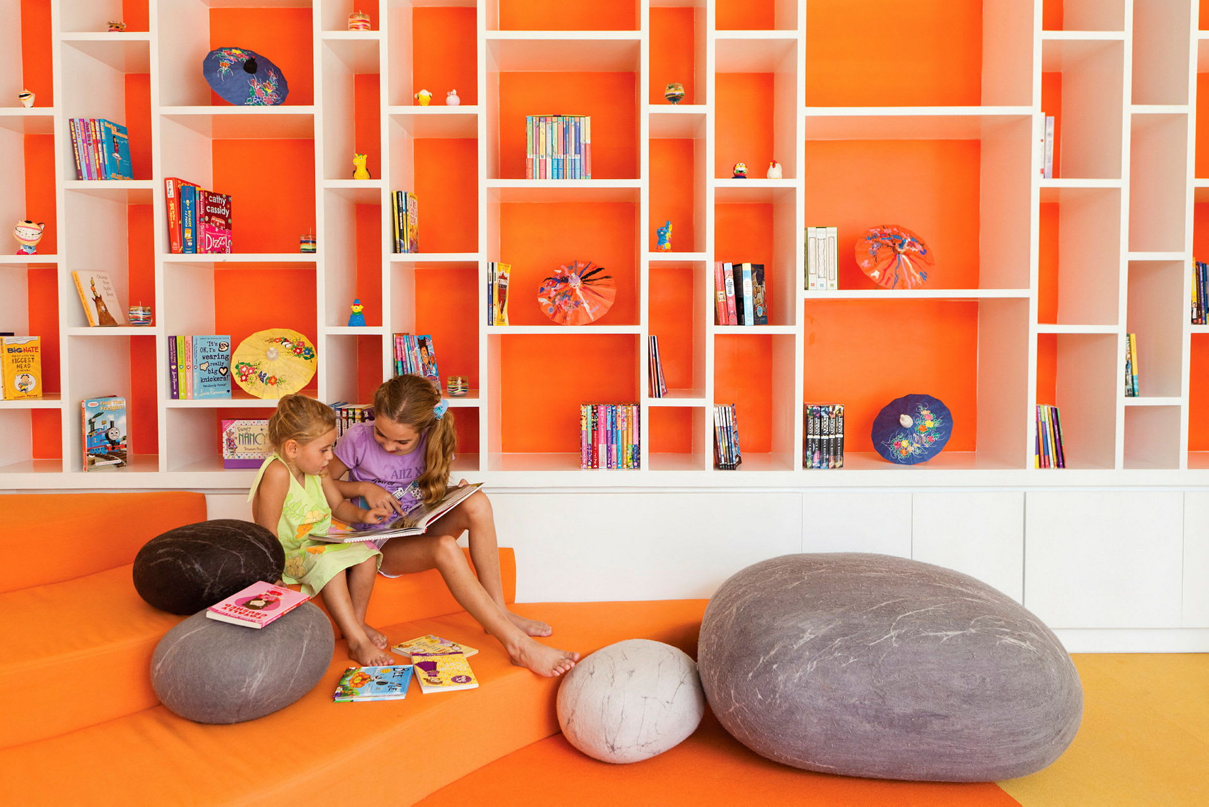 Två personer sitter och läser en bok på ett färgglatt bibliotek med orange bakgrund och hyllor fyllda med böcker och dekorationer. (AI-genererad text)