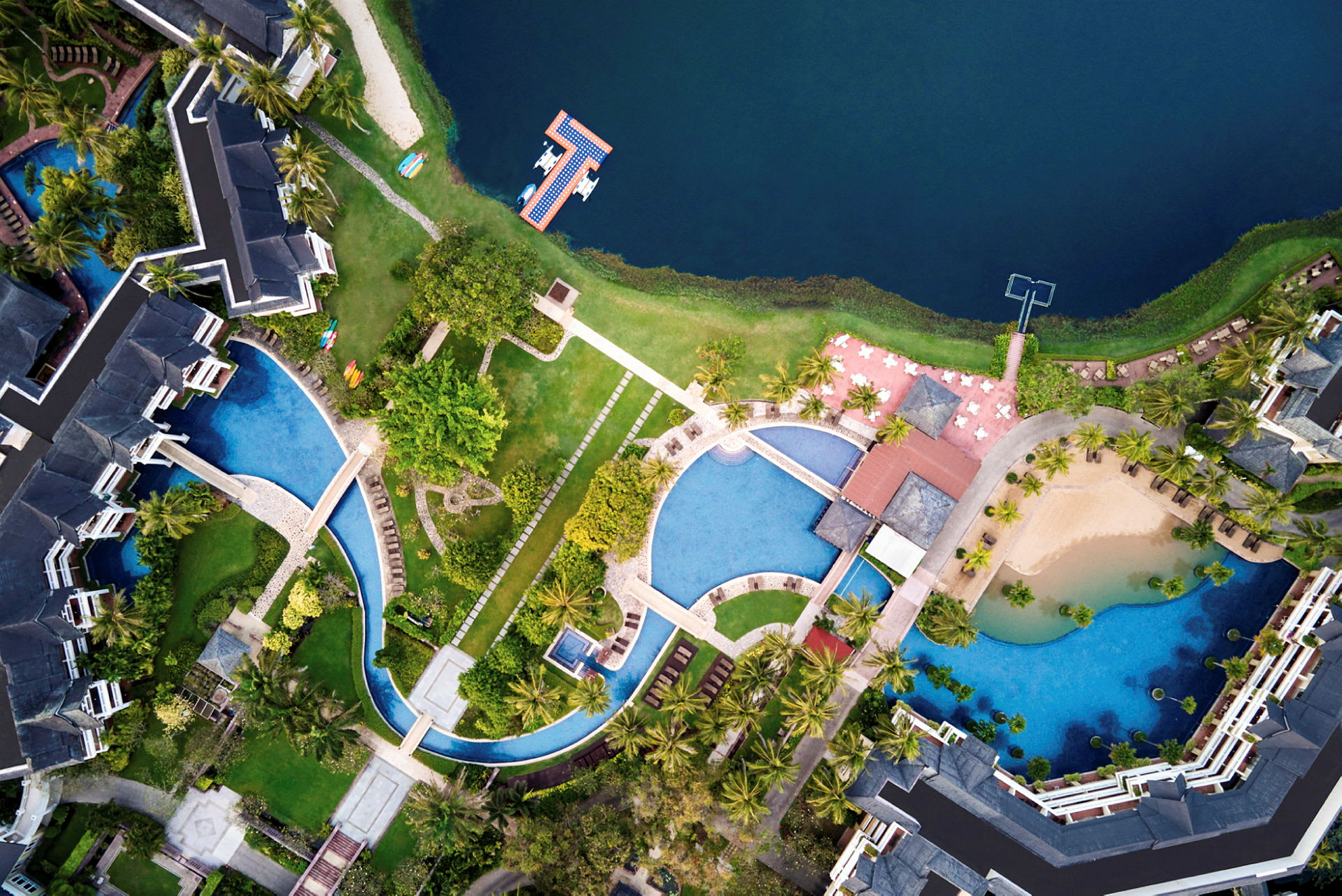 Flygfoto över hotellområde med byggnader, pooler, grönska och omgivande vatten. (AI-genererad text)