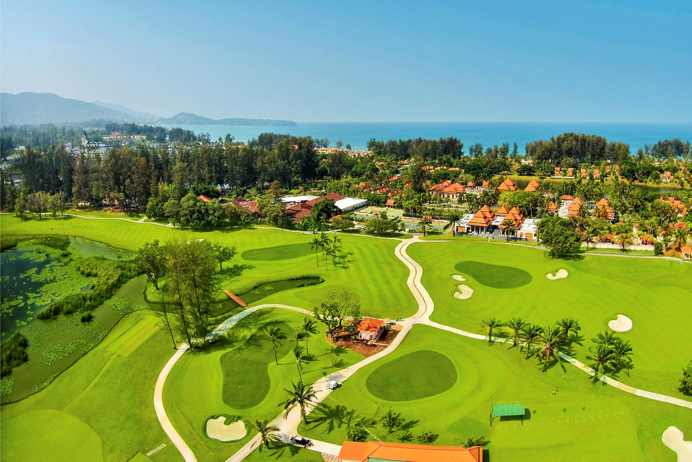 En golfbana med gröna fairways, bunkrar och vattenhinder, omgiven av lummiga träd och en resort nära havet. (AI-genererad text)
