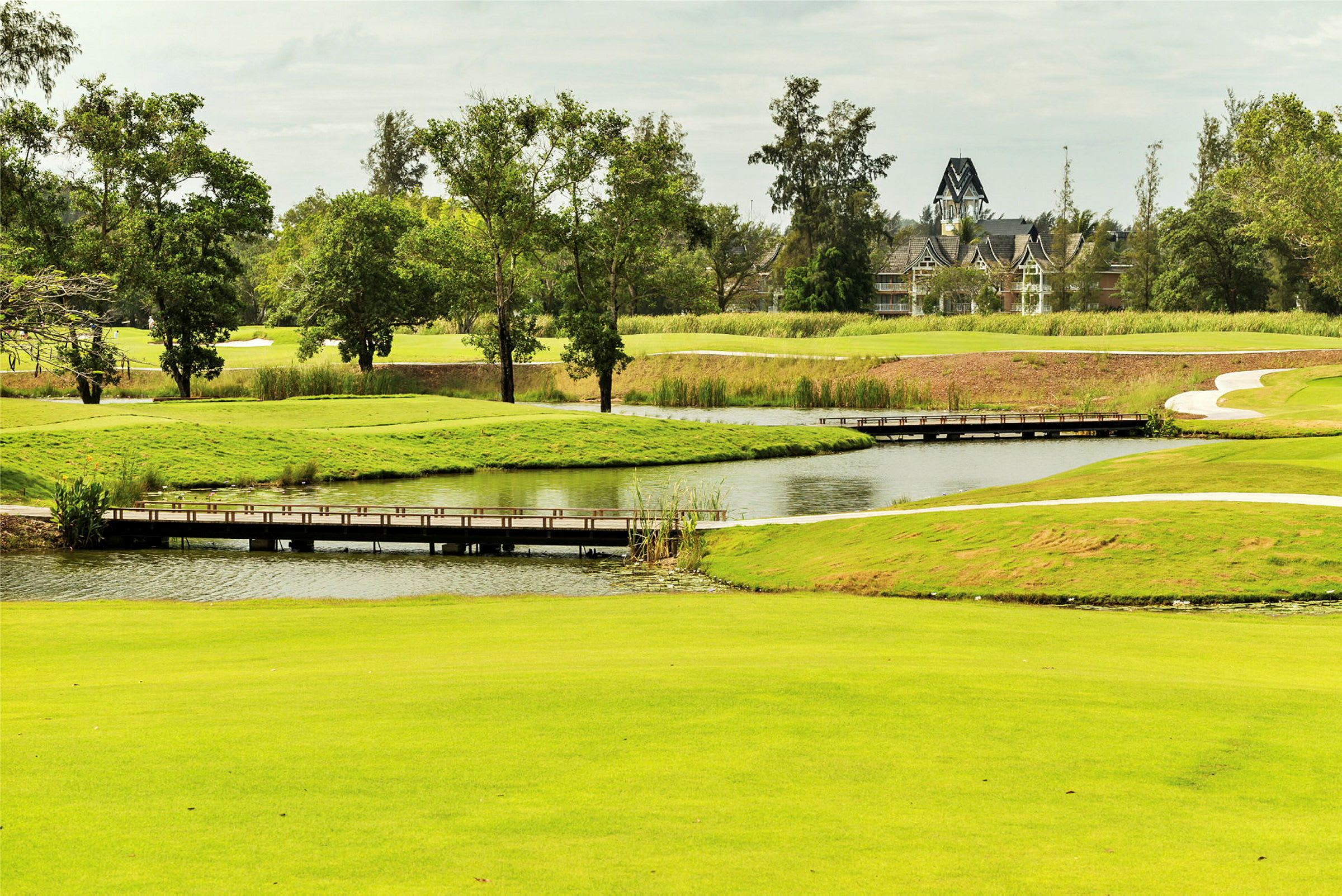En bild av en grön golfbana med en liten gångbro som går över vatten, omgiven av träd och vegetation. (AI-genererad text)