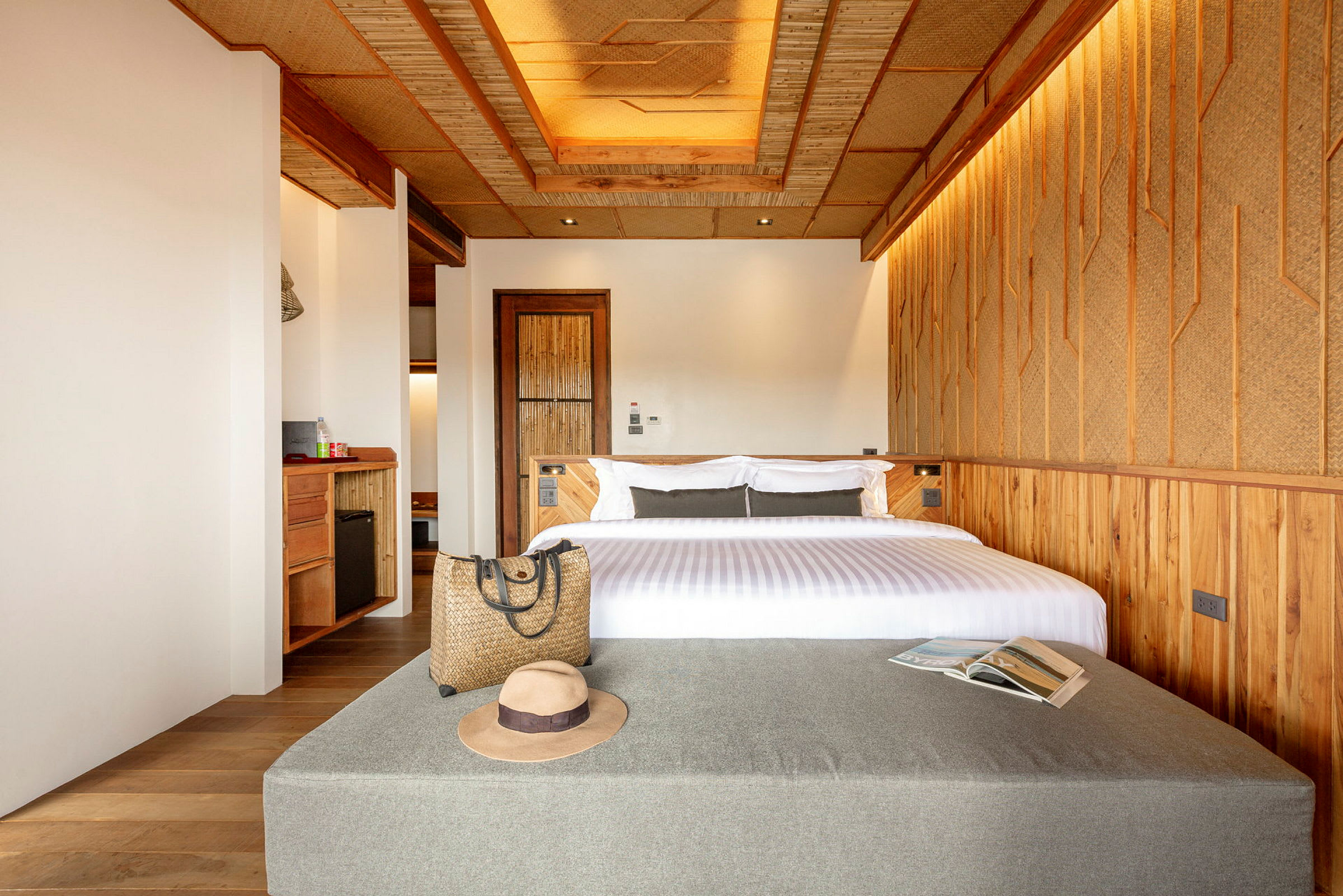 Ett hotellrum med en modern och varm inredning, där en dubbelsäng är placerad med kuddar och en vit bäddset. Framtill finns en grå bänk med en hatt, en väska och en öppen tidning ovanpå. Väggarna är dekorerade med trä och naturliga material. (AI-genererad text)