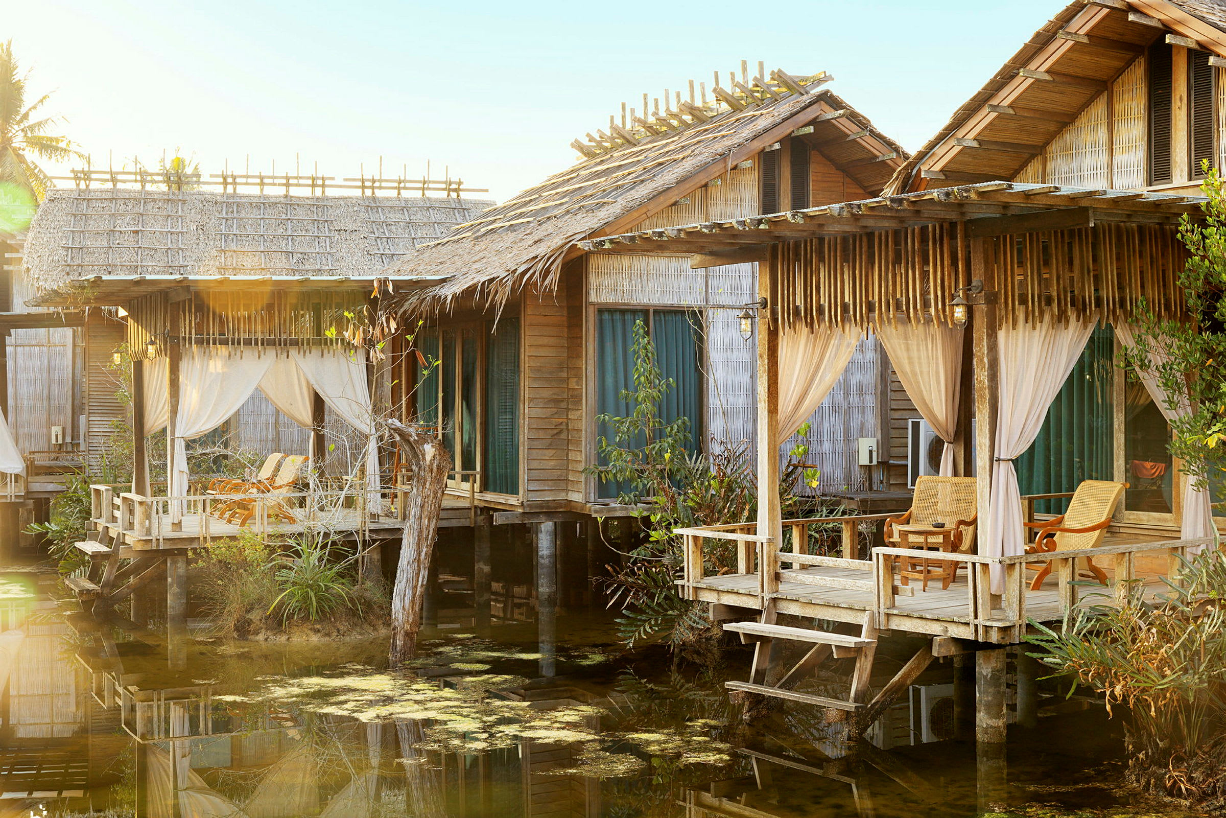 Vattennära bungalows med trästruktur och genomskinliga gardiner; verandor med sittmöbler. (AI-genererad text)