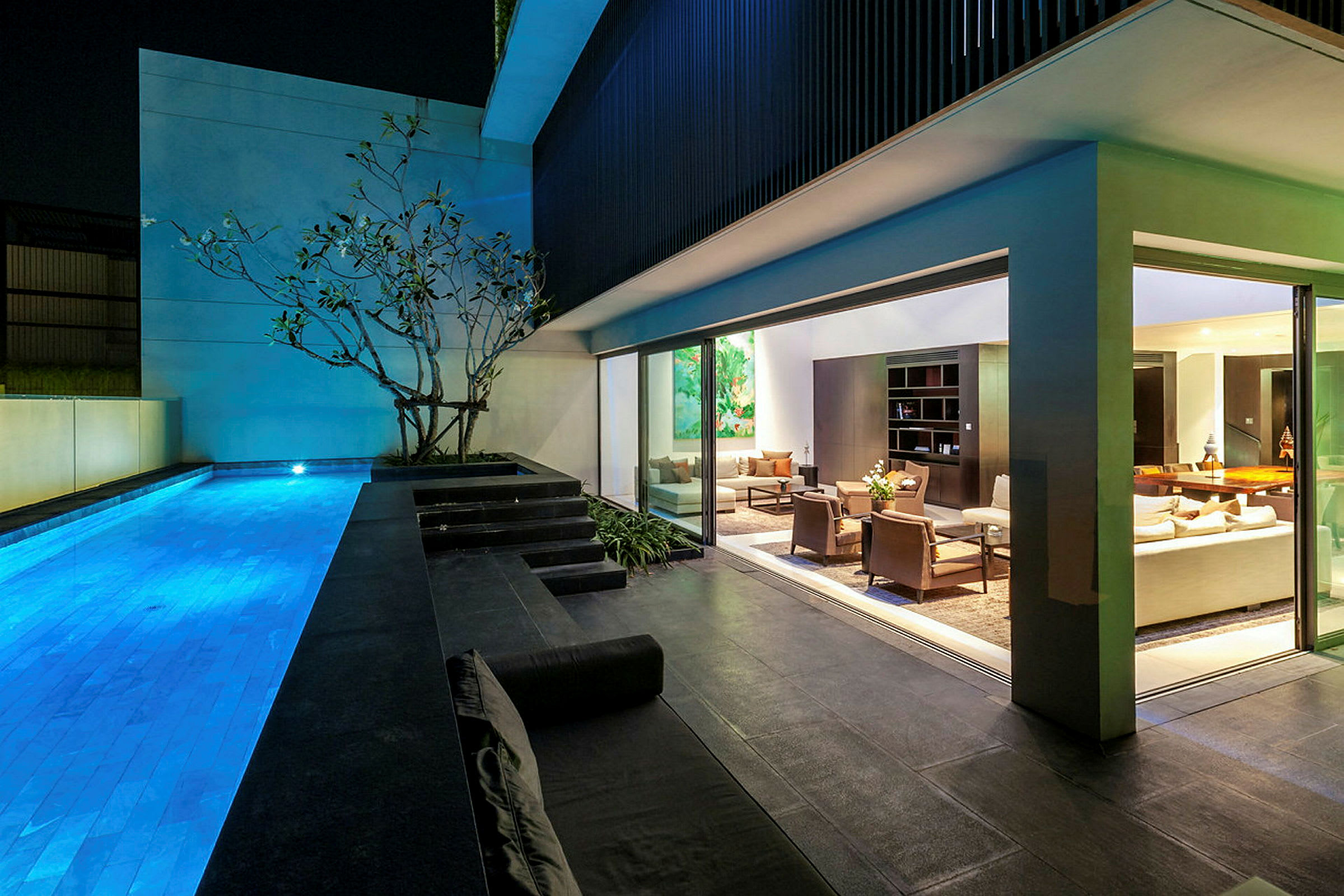 Bild av en modern villa med en upplyst swimmingpool utanför och ett bekvämt inrett vardagsrum på insidan synligt genom glasväggar. (AI-genererad text)