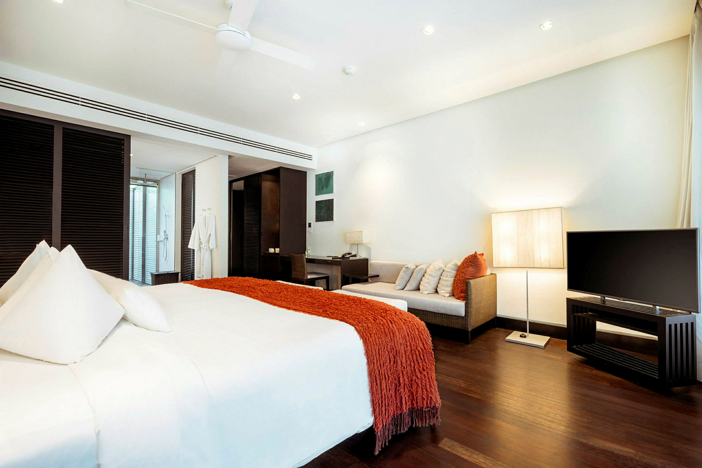 Ett modernt hotellrum med en stor säng klädd i vita lakan och en orange filt, en soffa med kuddar, en skrivbordshörna och en TV. Rummet har mörkt trägolv och är väl upplyst. (AI-genererad text)