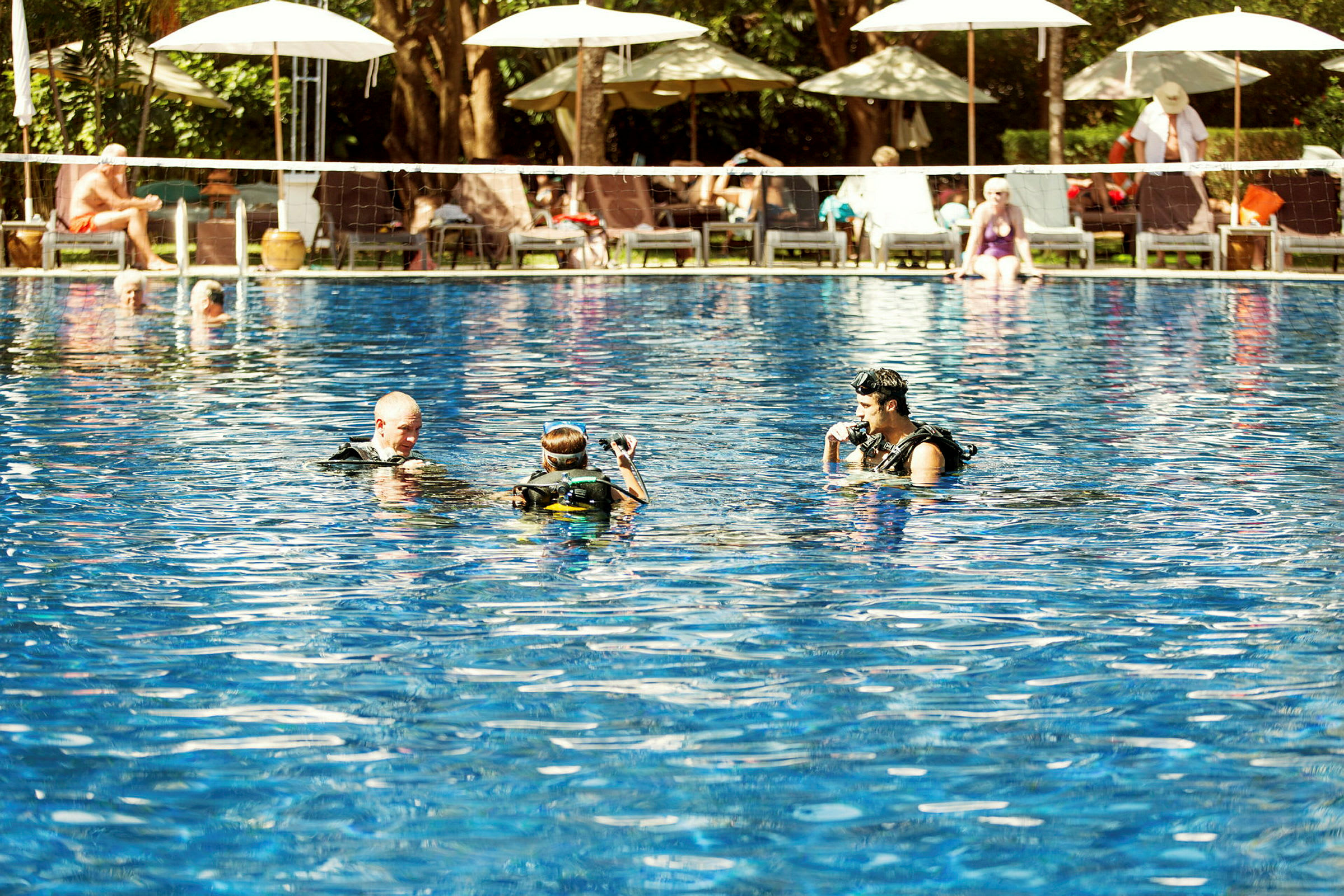 Personer i dykutrustning i en pool med solstolar och parasoller i bakgrunden. (AI-genererad text)