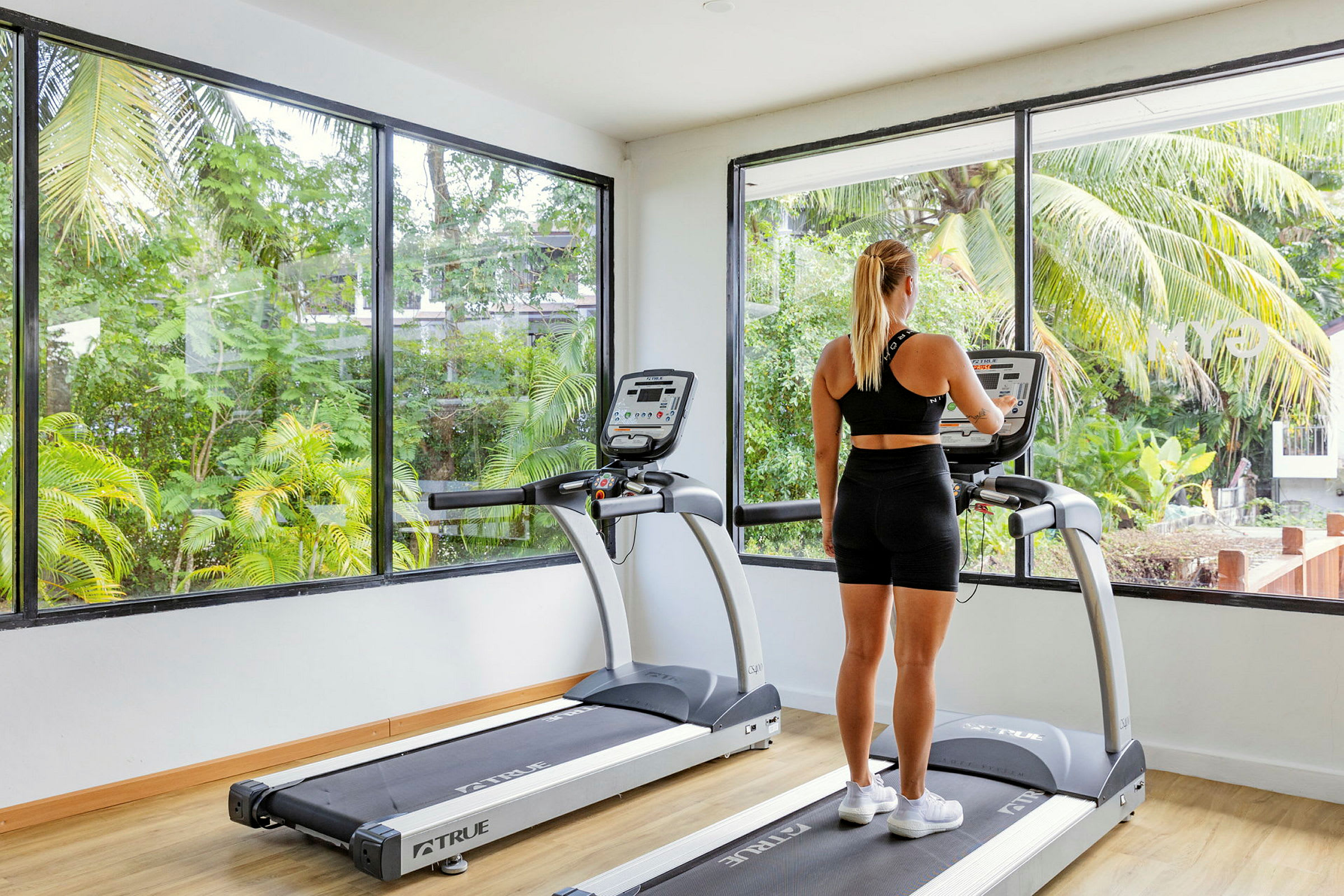 En person tittar på skärmen av en löpmaskin i ett gym med stora fönster och utsikt över grönska. (AI-genererad text)
