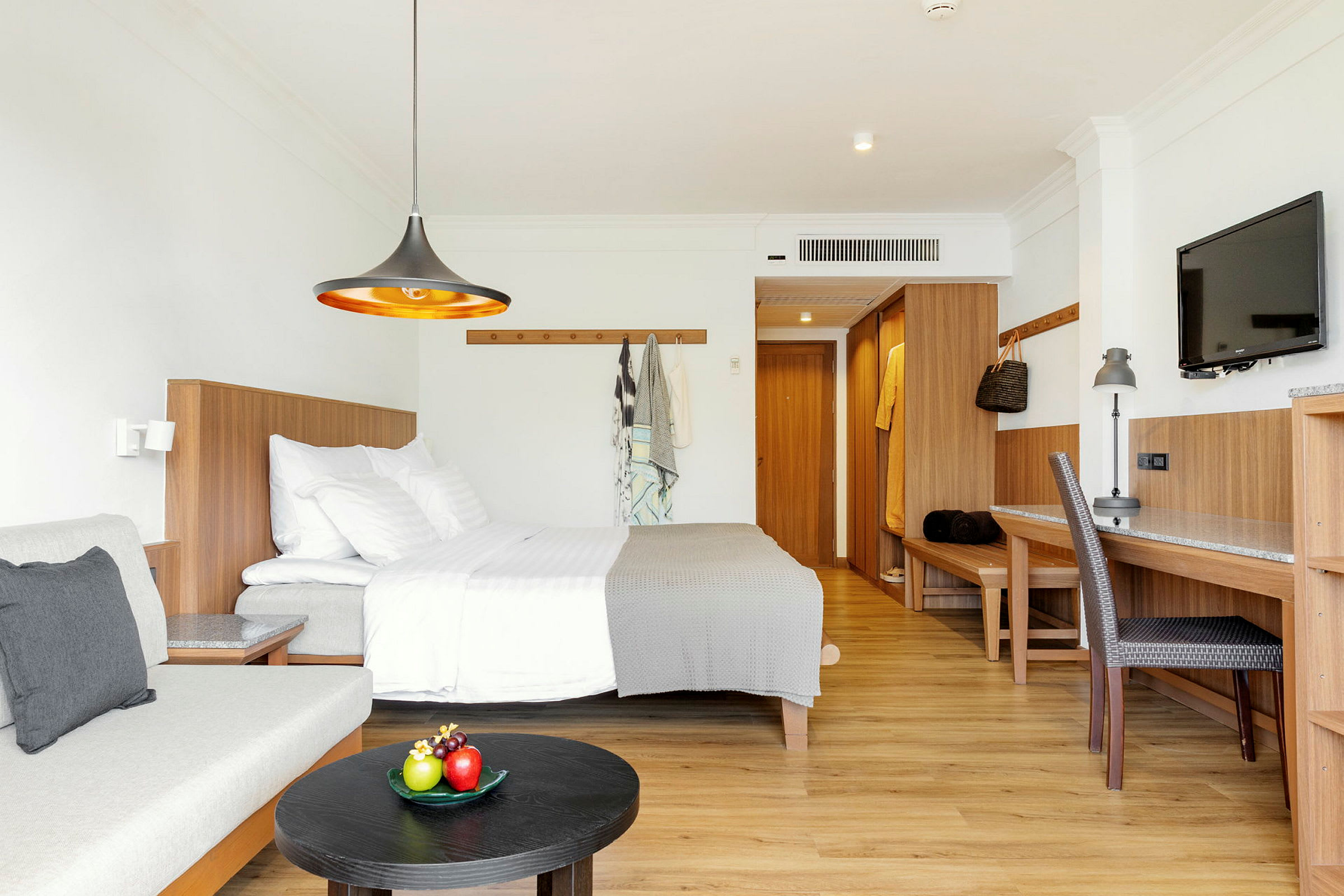 Ett modernt hotellrum med säng, soffa, skrivbord och en TV, inrett i ljusa färger och trädetaljer. (AI-genererad text)