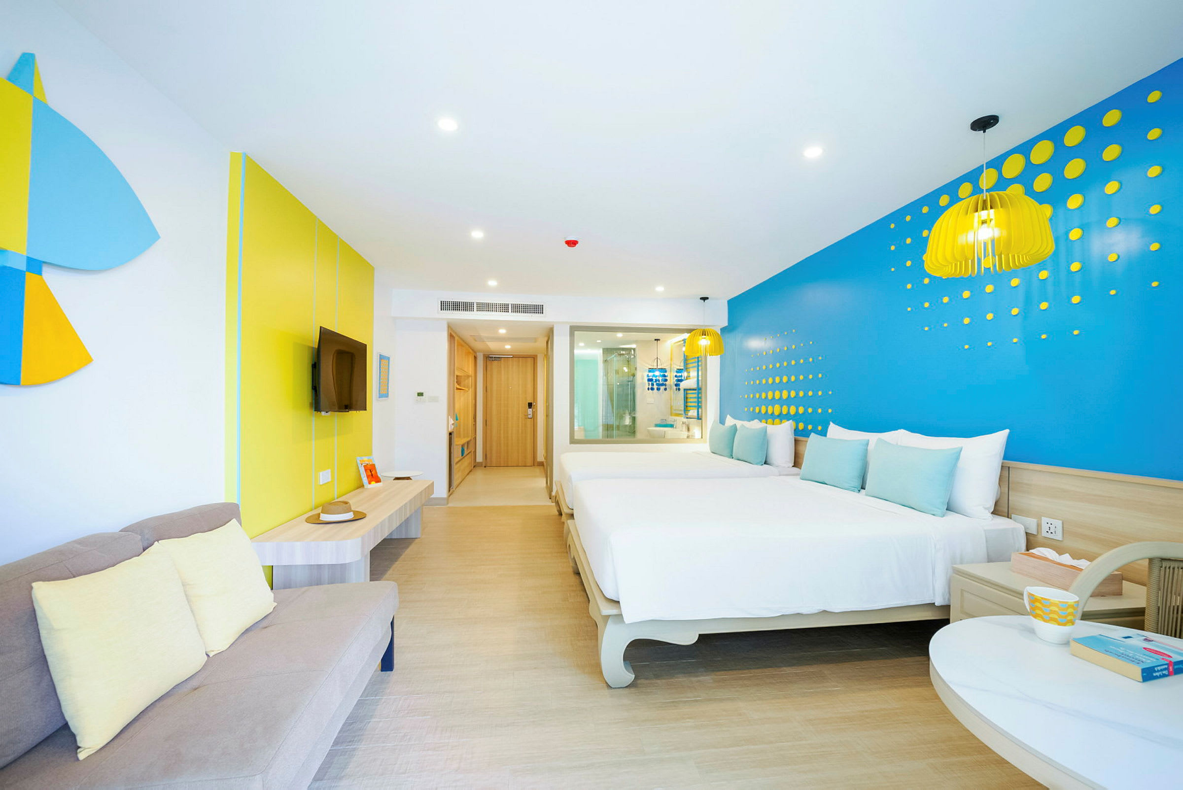 Ett ljust hotellrum med en modern inredning i blått och gult, en säng, en soffa och ett tv-område. (AI-genererad text)