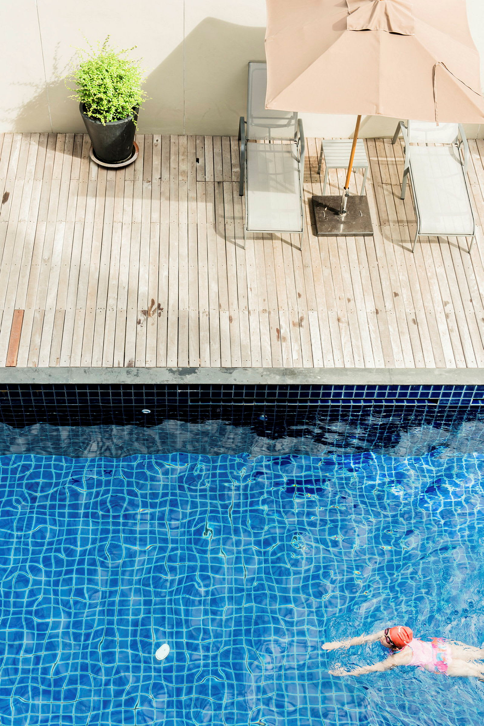 En person simmar i en pool med blått kakel. Vid sidan av poolen finns en träterrass med solstolar, ett parasoll och en krukväxt. (AI-genererad text)