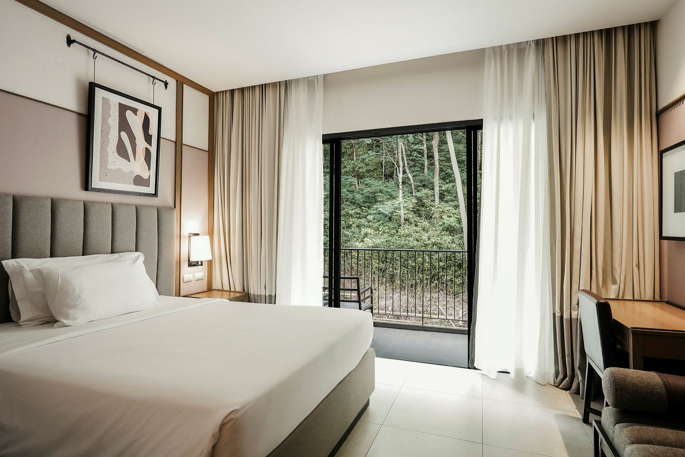 Ett hotellrum med en stor säng, väggdekoration, skrivbord och stol. Glasdörrar leder ut till en balkong med utsikt över ett grönområde. (AI-genererad text)
