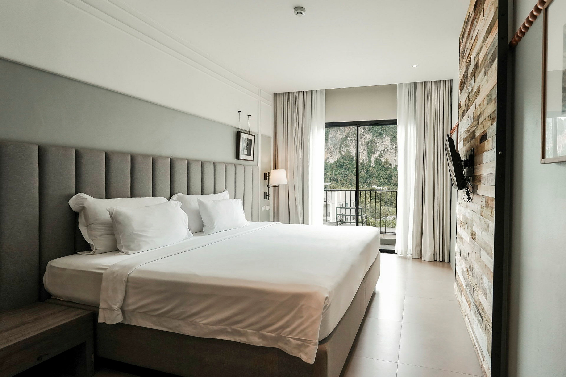 Ett hotellrum med en stor dubbelsäng, modern inredning och en glasdörr som vetter mot en balkong med utsikt över gröna träd och berg. (AI-genererad text)