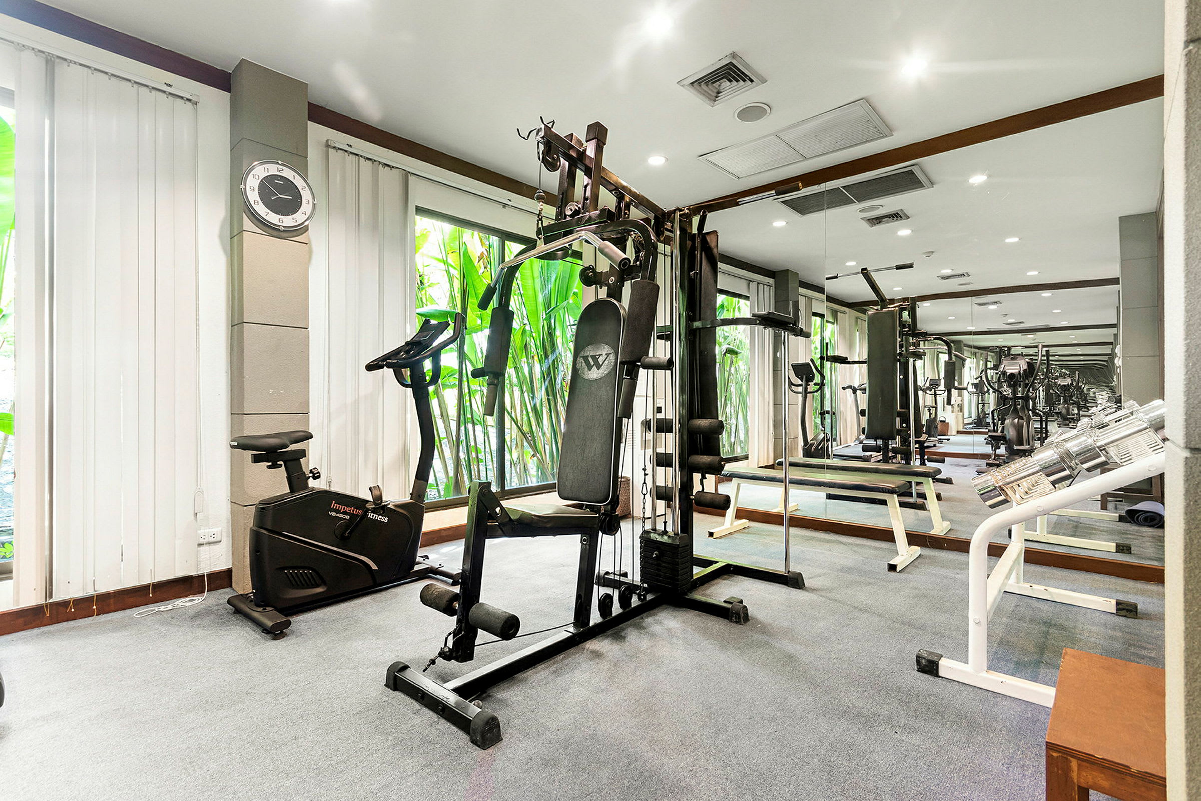 Ett gym med träningsmaskiner och vikter som står i ett rum med spegelväggar och fönster som släpper in dagsljus. (AI-genererad text)