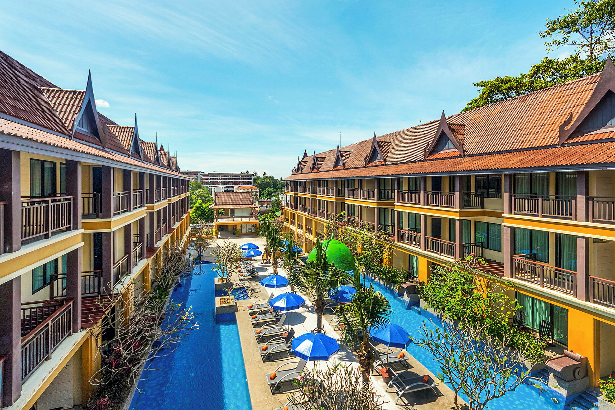 Två våningar höga hotellbyggnader med sluttande tak omger en pool med solstolar och parasoller. (AI-genererad text)