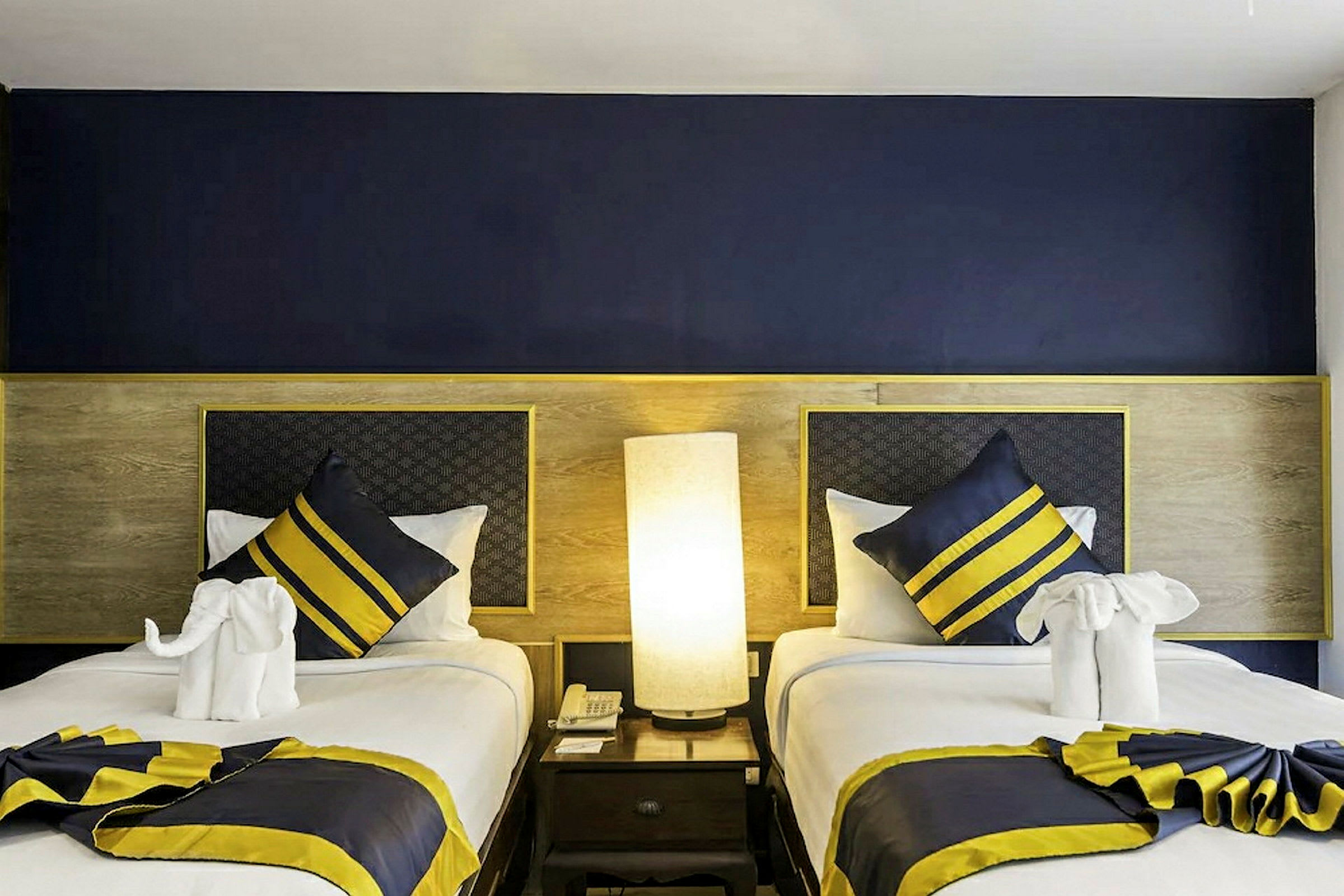 Ett hotellrum med två enkelsängar, prydda med dekorativa kuddar och vikta handdukar. En lampa står på bordet mellan sängarna. (AI-genererad text)