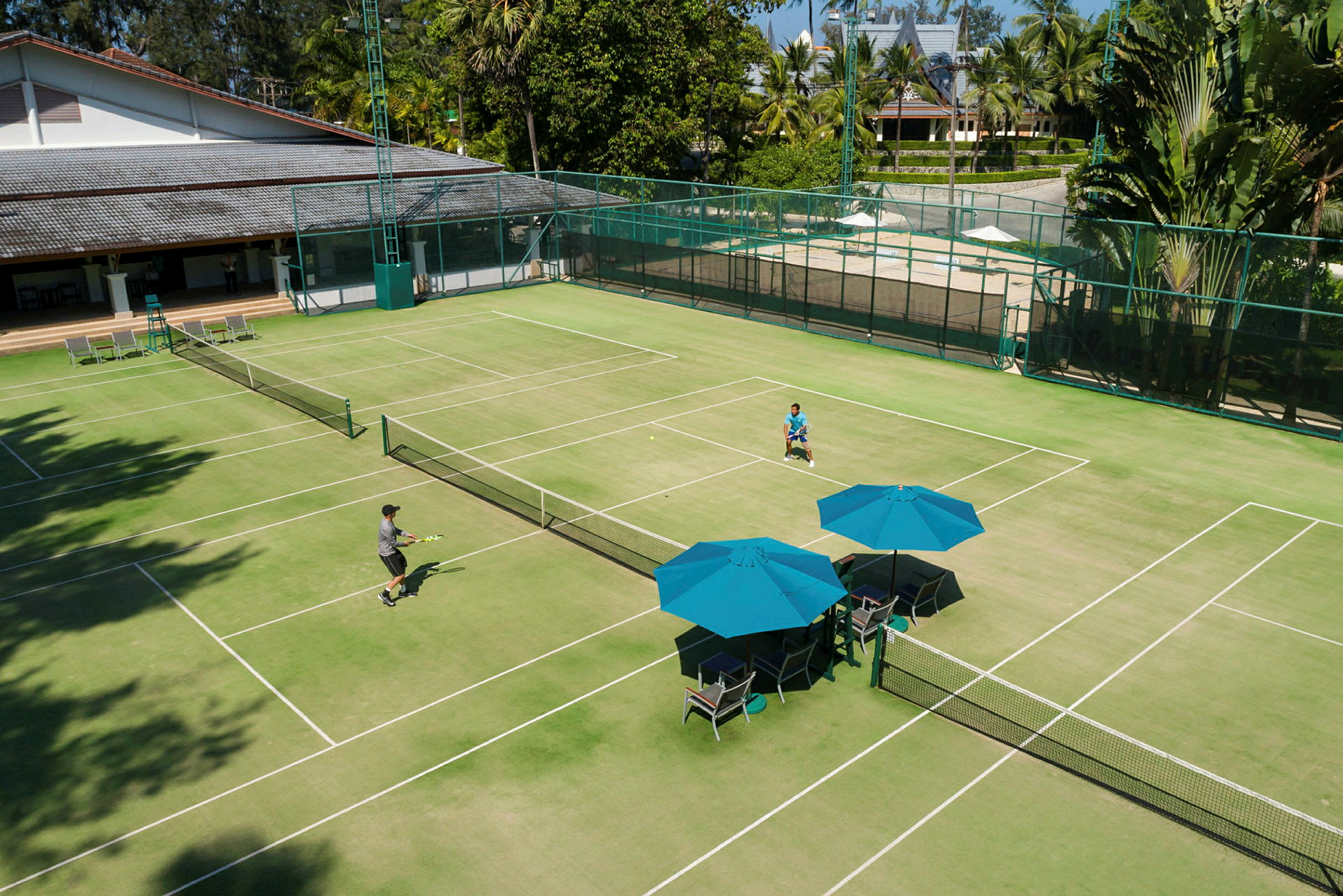 En grön tennisbana med två personer som spelar tennis. Det finns blå parasoller och stolar vid sidan av banan. (AI-genererad text)