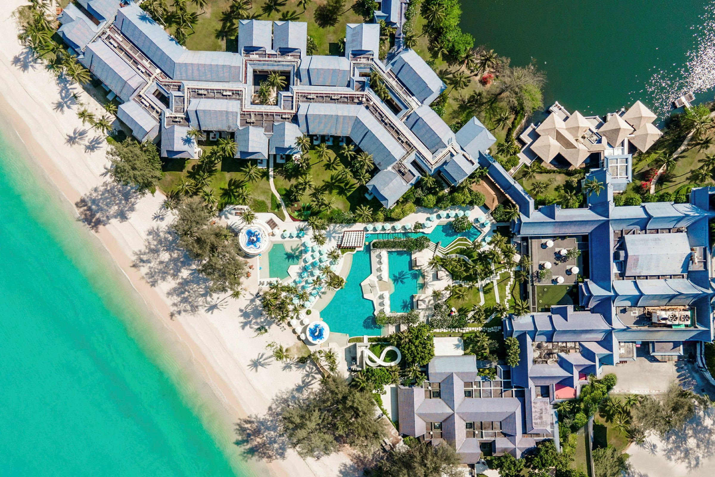 Flygbild över en resort med poolområden och palmer vid en strand. (AI-genererad text)