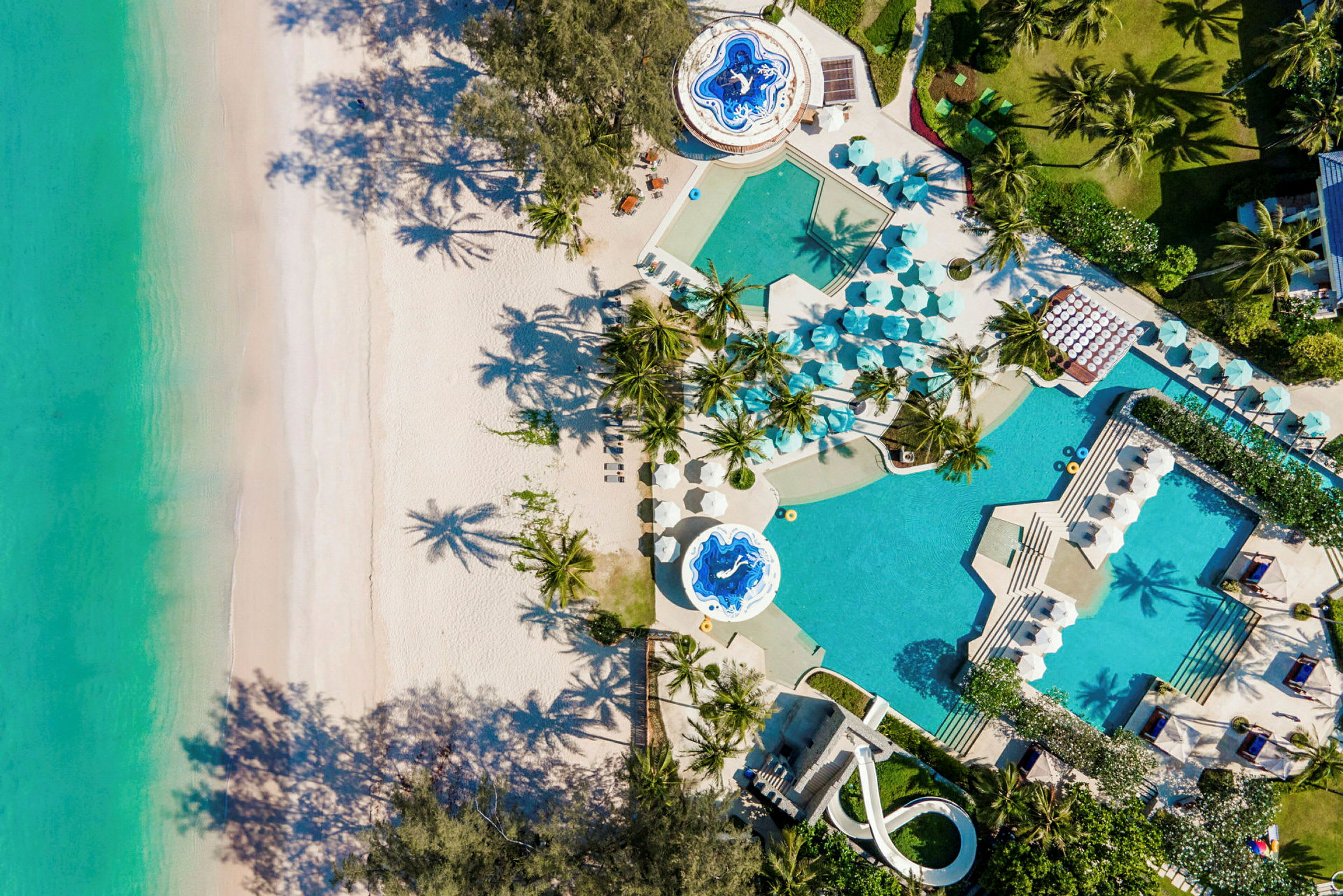 Flygfoto över en strand med vit sand och klart turkost vatten. Intill stranden finns ett poolområde med parasoller, växter och palmer. (AI-genererad text)