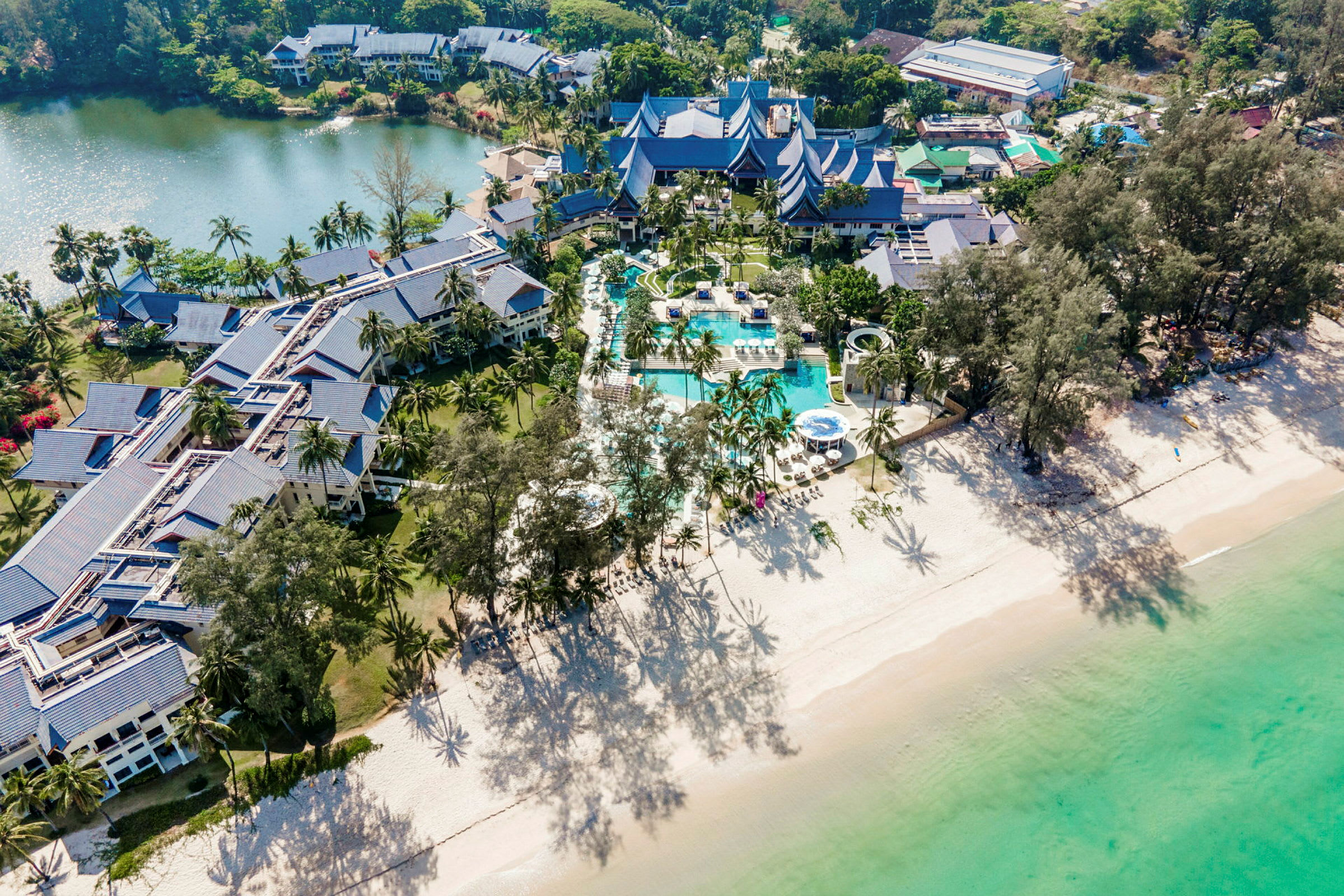 Flygbild av ett hotellkomplex vid en sandstrand och grönt vatten, omgiven av grön natur och palmer. (AI-genererad text)