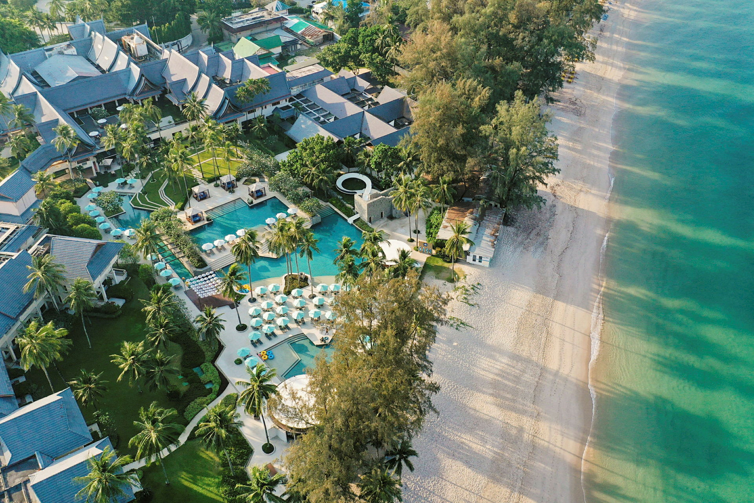 Flygfoto över en resort med pool och omgivning av palmträd vid en sandstrand. (AI-genererad text)