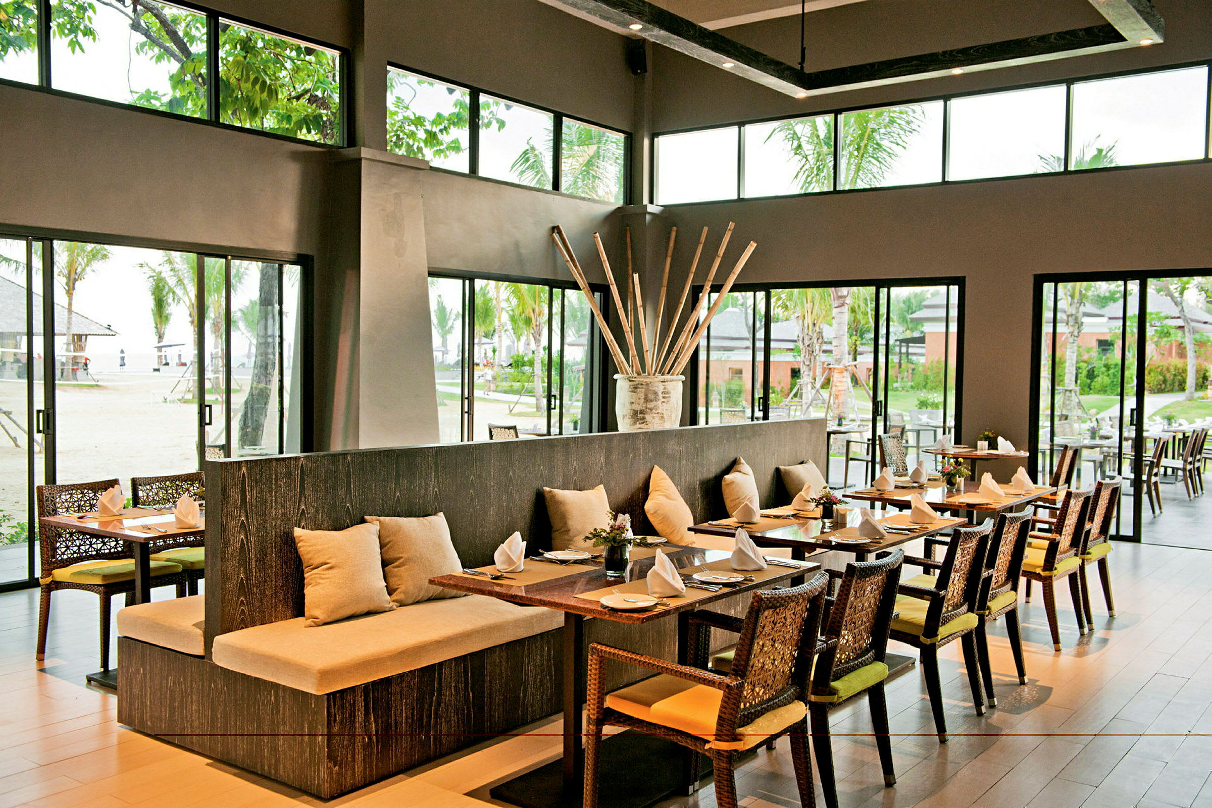Bild på en rymlig restaurang med stora fönster som vetter mot en utomhusmiljö med grönska och palmer. (AI-genererad text)