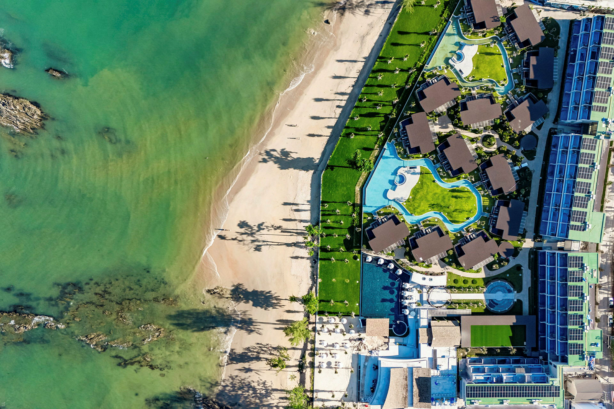 Flygbild av en strand och ett hotellområde med pooler och byggnader. (AI-genererad text)