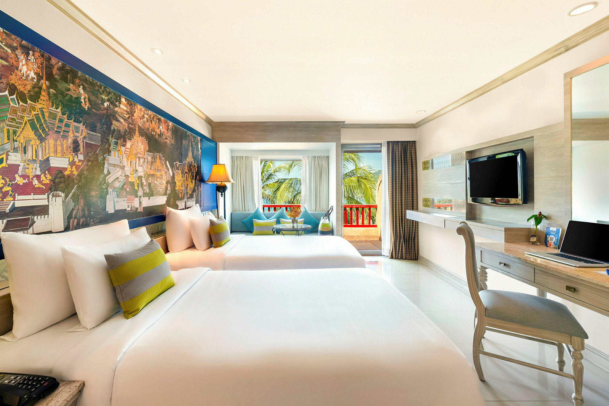 Ett hotellrum med två stora sängar, färgstarka kuddar och dekorativ målning på väggen. Fönster leder ut till en balkong med utsikt över palmer. (AI-genererad text)