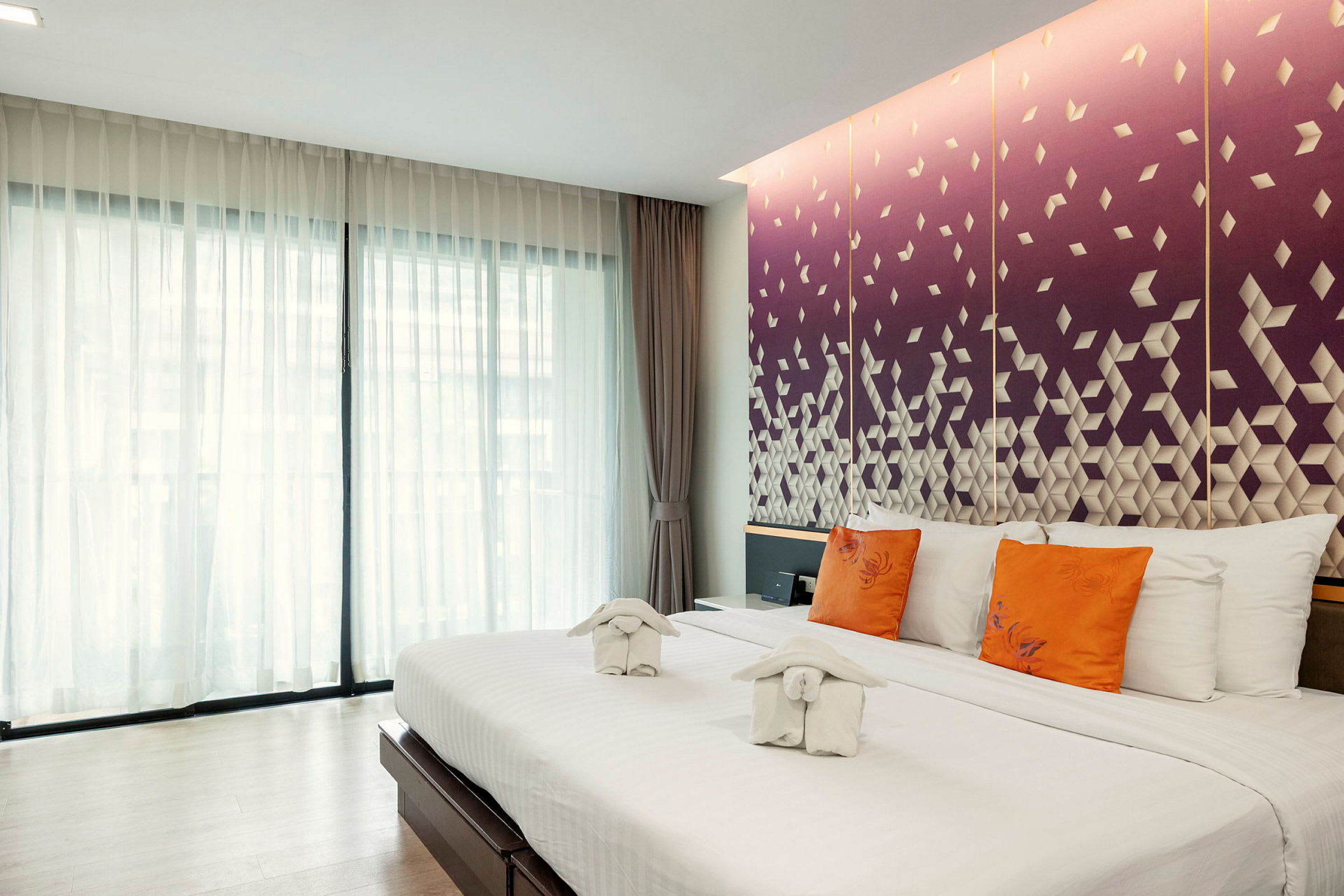 Ett hotellrum med en stor säng, vita sängkläder, orange kuddar och dekorativa handdukar. Bakgrunden visar en lila vägg med geometriskt mönster. (AI-genererad text)