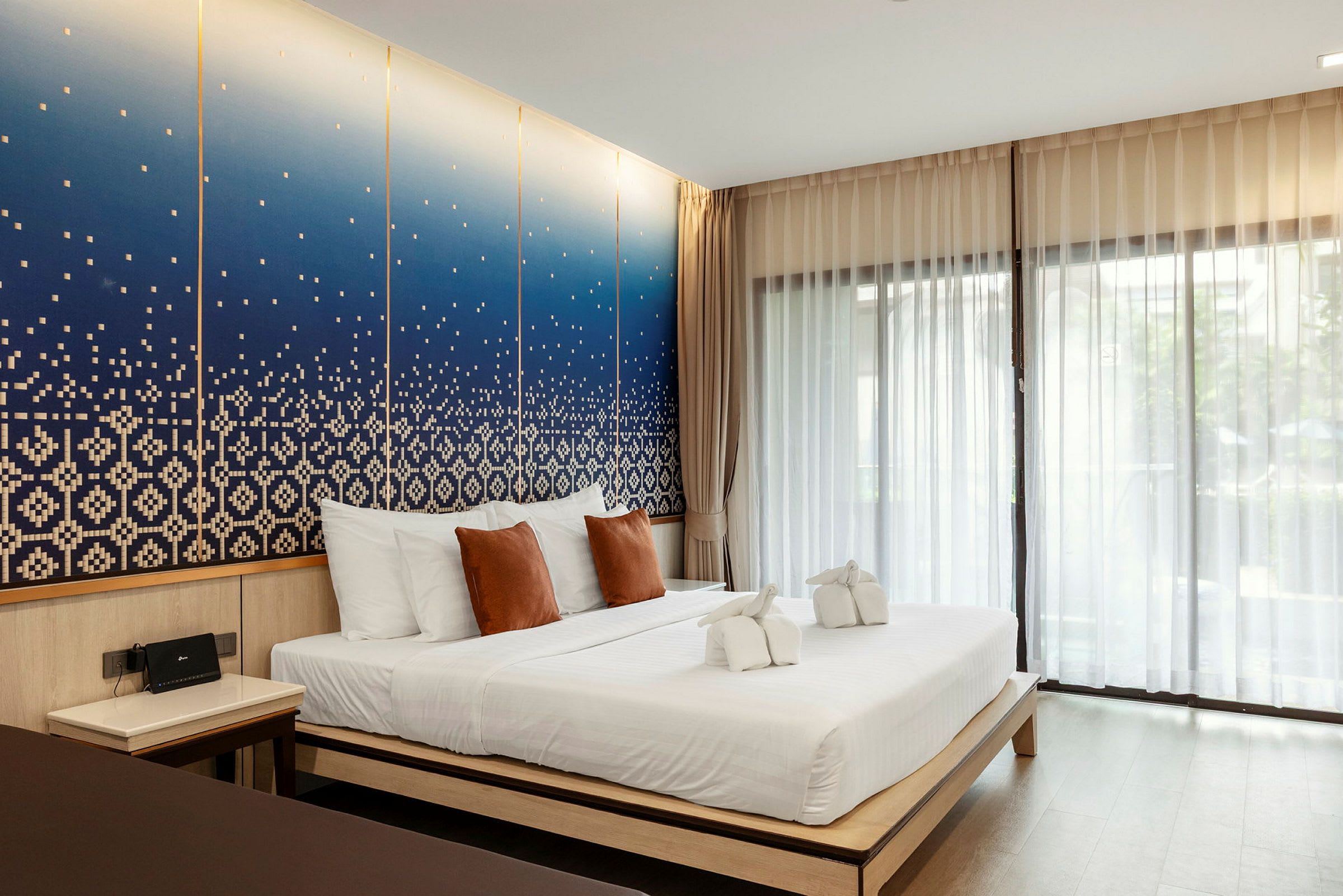 Ett hotellrum med en dubbelsäng, vita och bruna kuddar samt dekorativa handdukar på sängen. Bakgrunden har en blå konstnärlig väggdesign. (AI-genererad text)