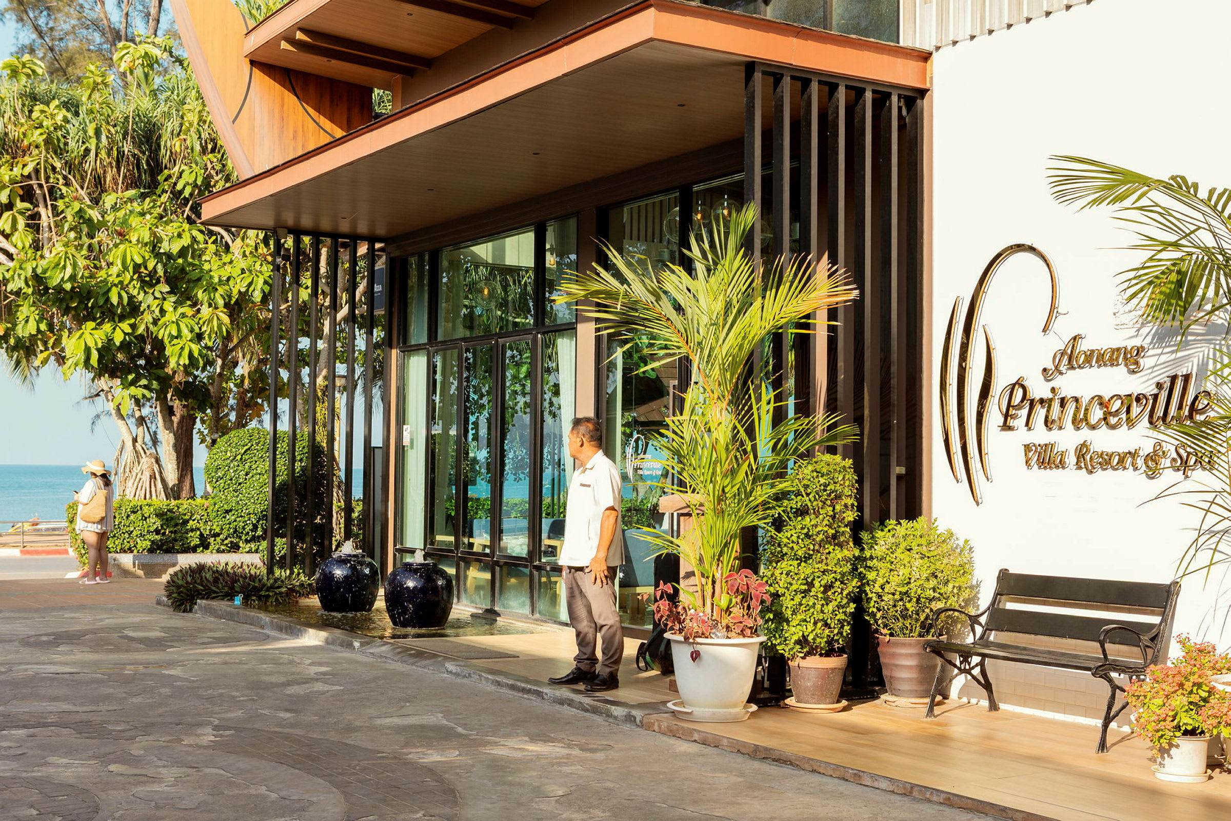 En entré till en resort med skylt som lyder "Aonang Princeville Villa Resort & Spa". Utomhus är planteringar och en bänk synliga, med bakgrund av hav och grönska. (AI-genererad text)