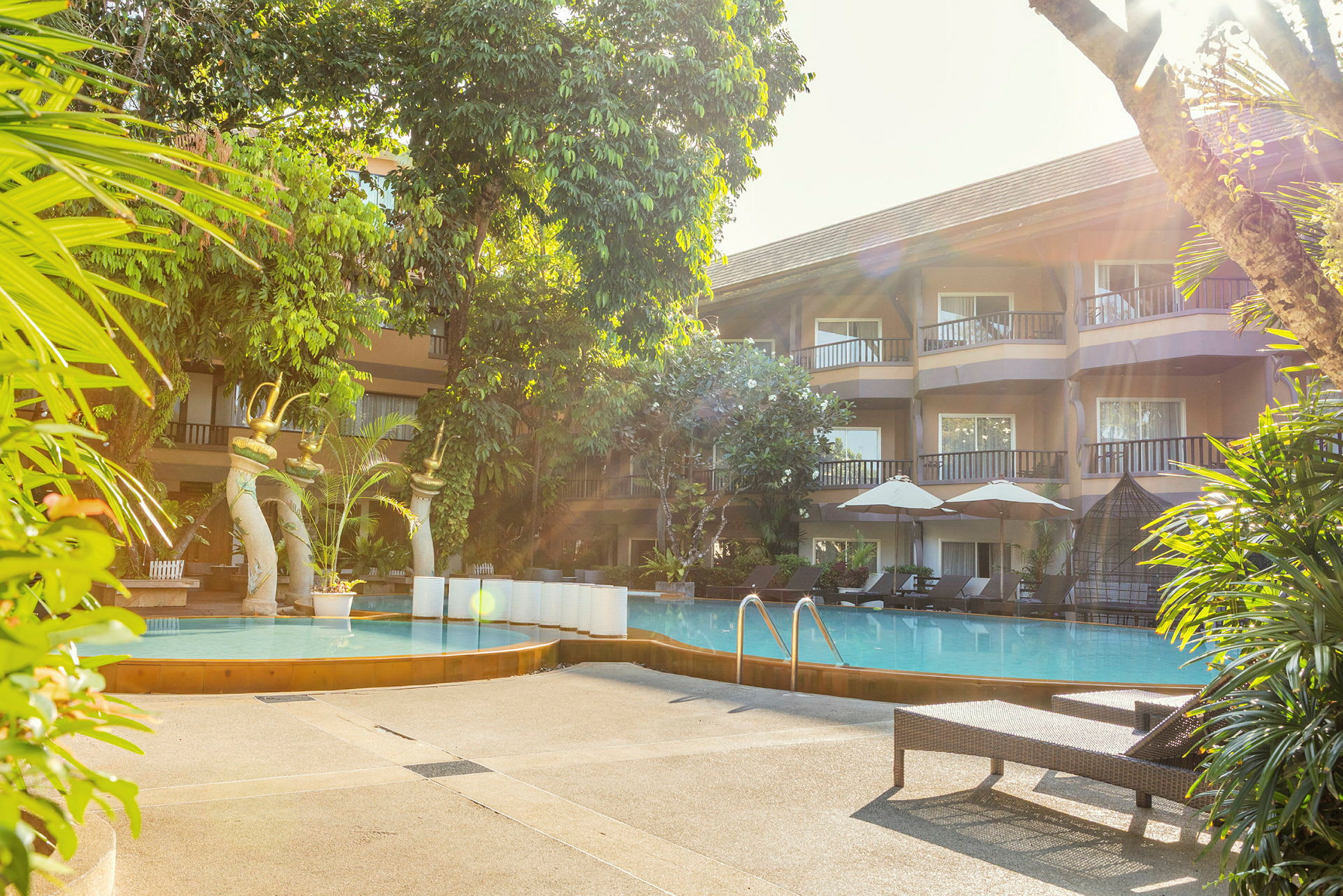Bild på en hotellpool omgiven av solstolar och parasoller, med frodig grönska och byggnaden i bakgrunden. (AI-genererad text)