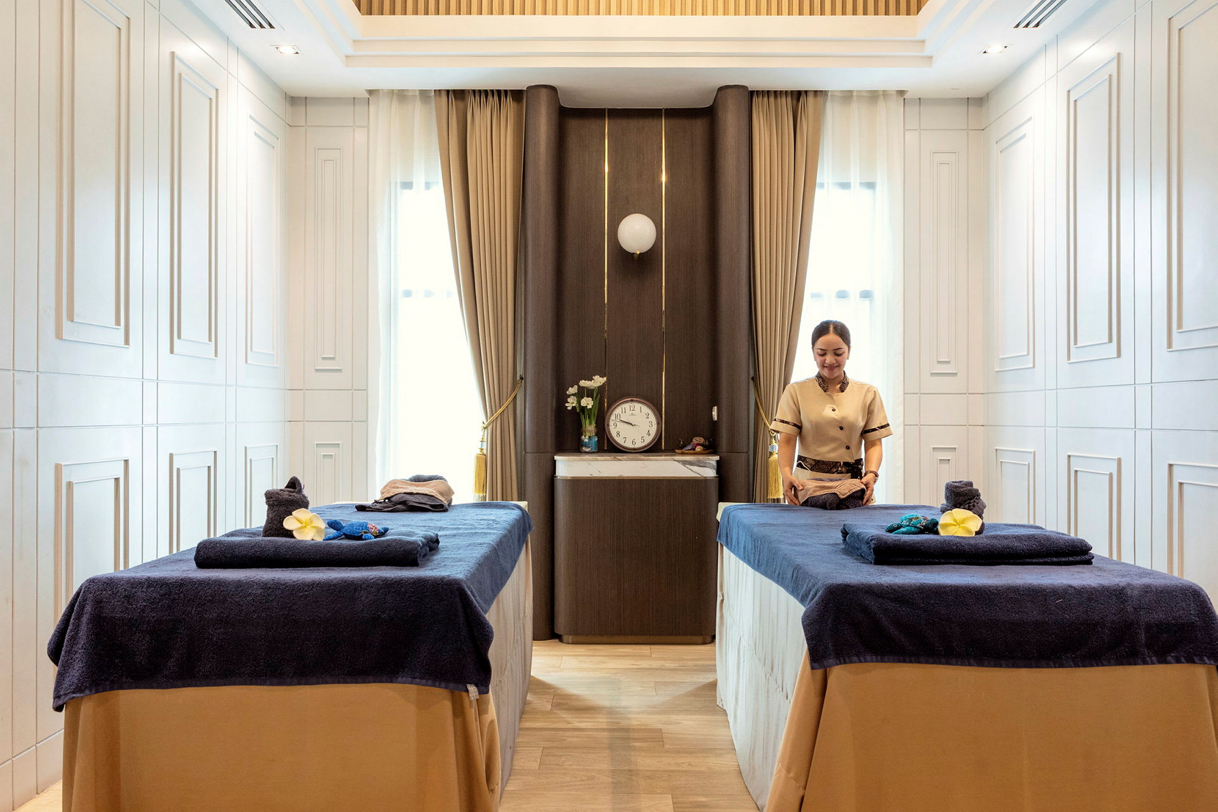 Bild av ett spa-rum med två massagebänkar klädda i mörkblå handdukar och en person som förbereder behandlingar. (AI-genererad text)