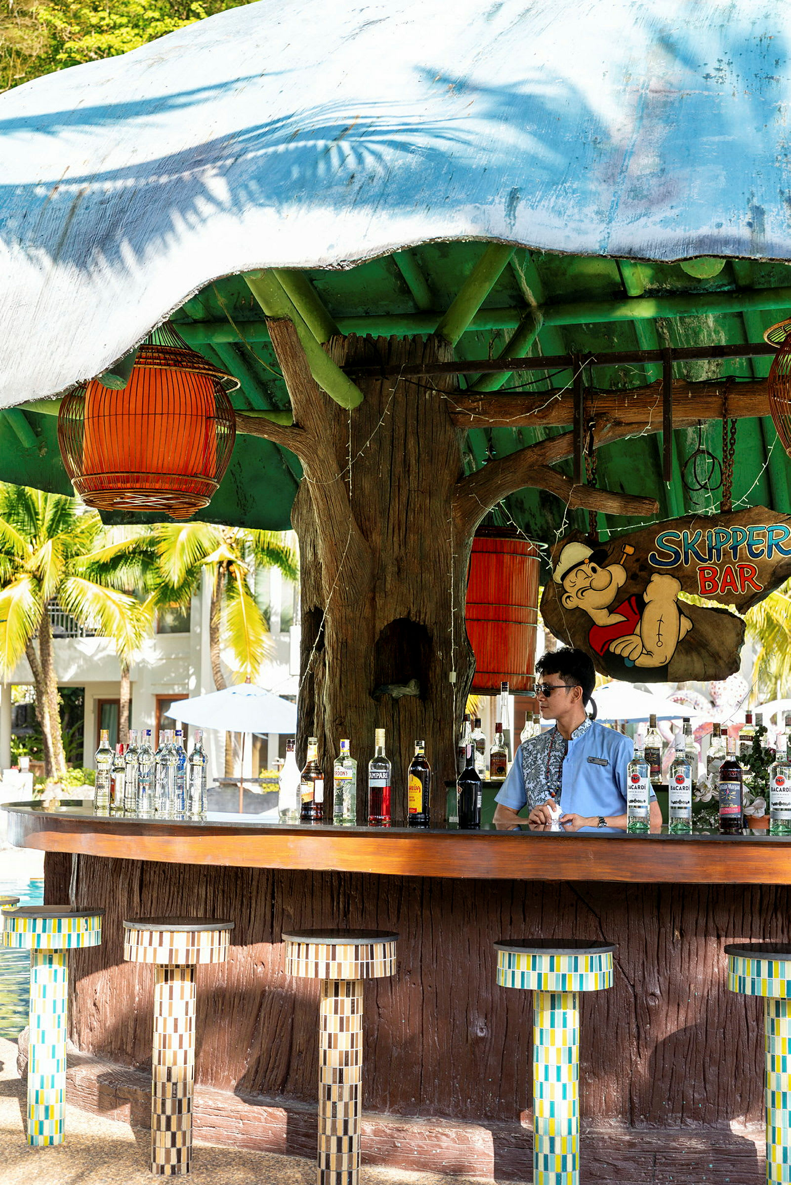En strandbar med färgglada stolar och olika spritflaskor på bardisken. En bartender står bakom baren. (AI-genererad text)