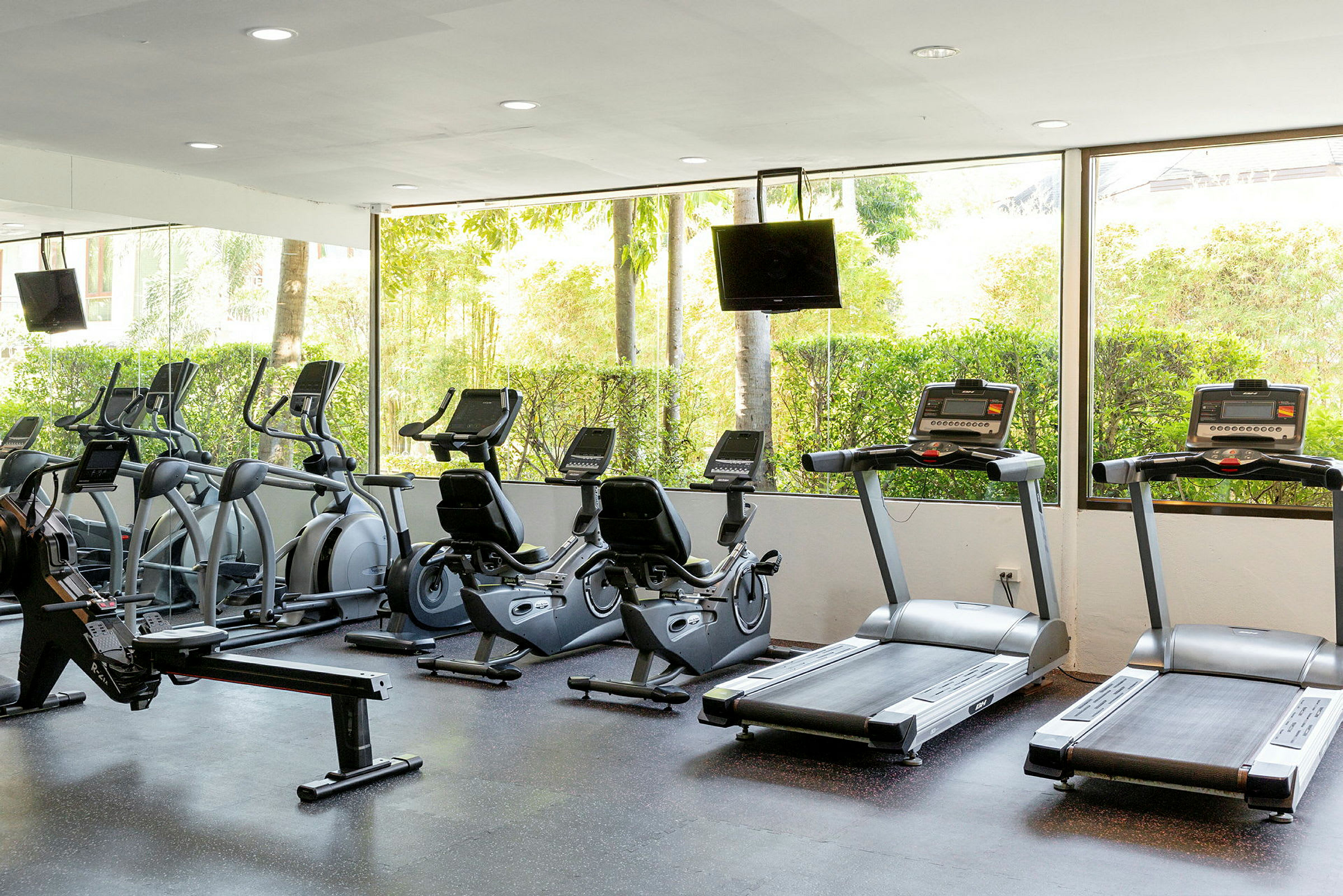 Ett gym med löpband, träningscyklar och träningsutrustning framför stora fönster med utsikt mot grönska. (AI-genererad text)