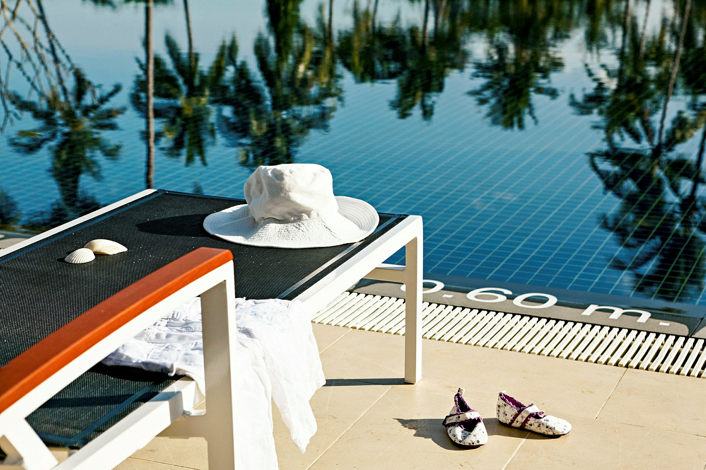 Solhatt och snäckskal på solstol vid pool. Reflektion av palmer i vattnet. (AI-genererad text)