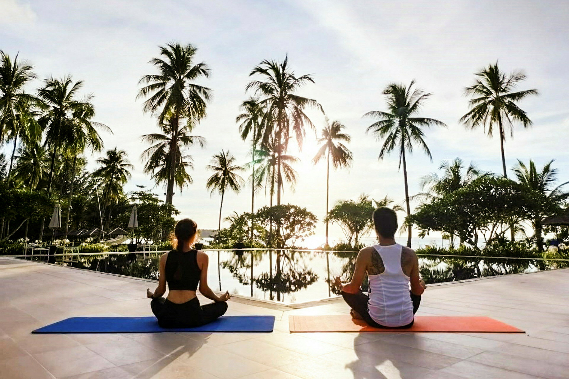 Två personer sitter på yogamattor vid en pool omgiven av palmer. (AI-genererad text)
