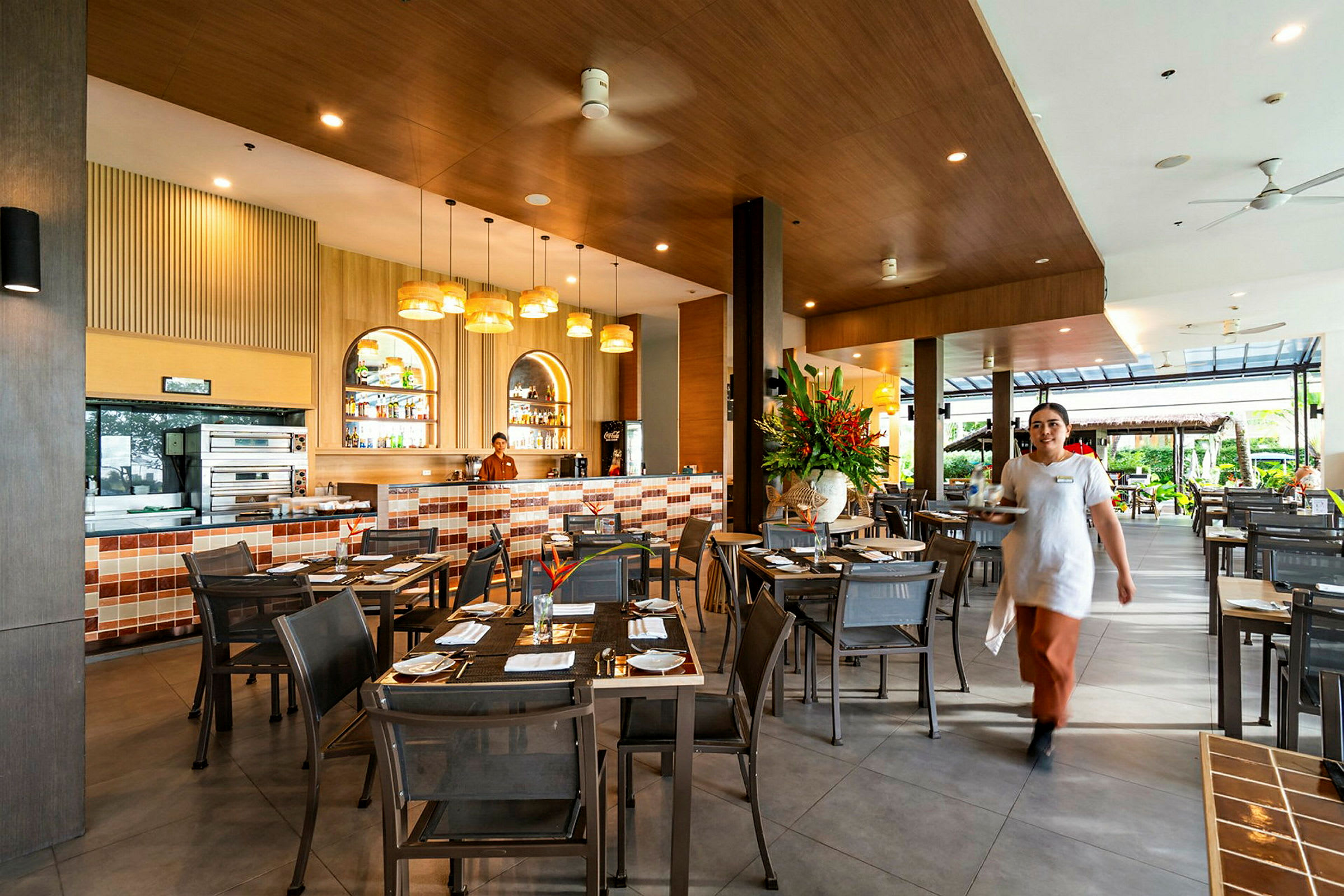 En restaurang med bord och stolar; i bakgrunden syns en bar med kaklade detaljer och en anställd. En serveringspersonal går mot kameran. (AI-genererad text)