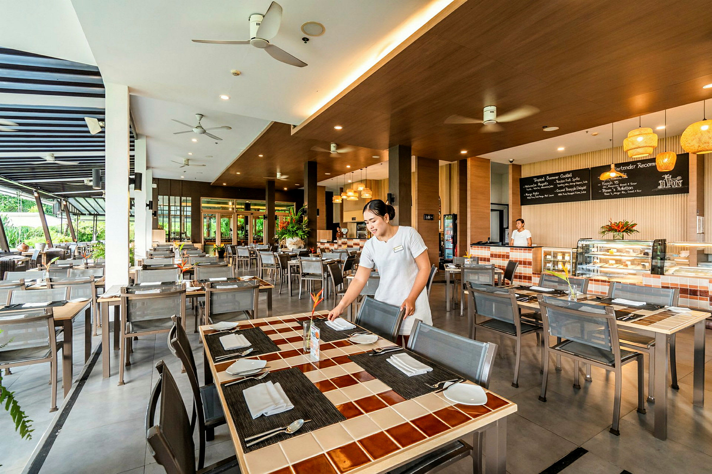 En ljus restaurang med moderna möbler, en medarbetare arrangerar bord. Stor bardisk syns i bakgrunden. (AI-genererad text)