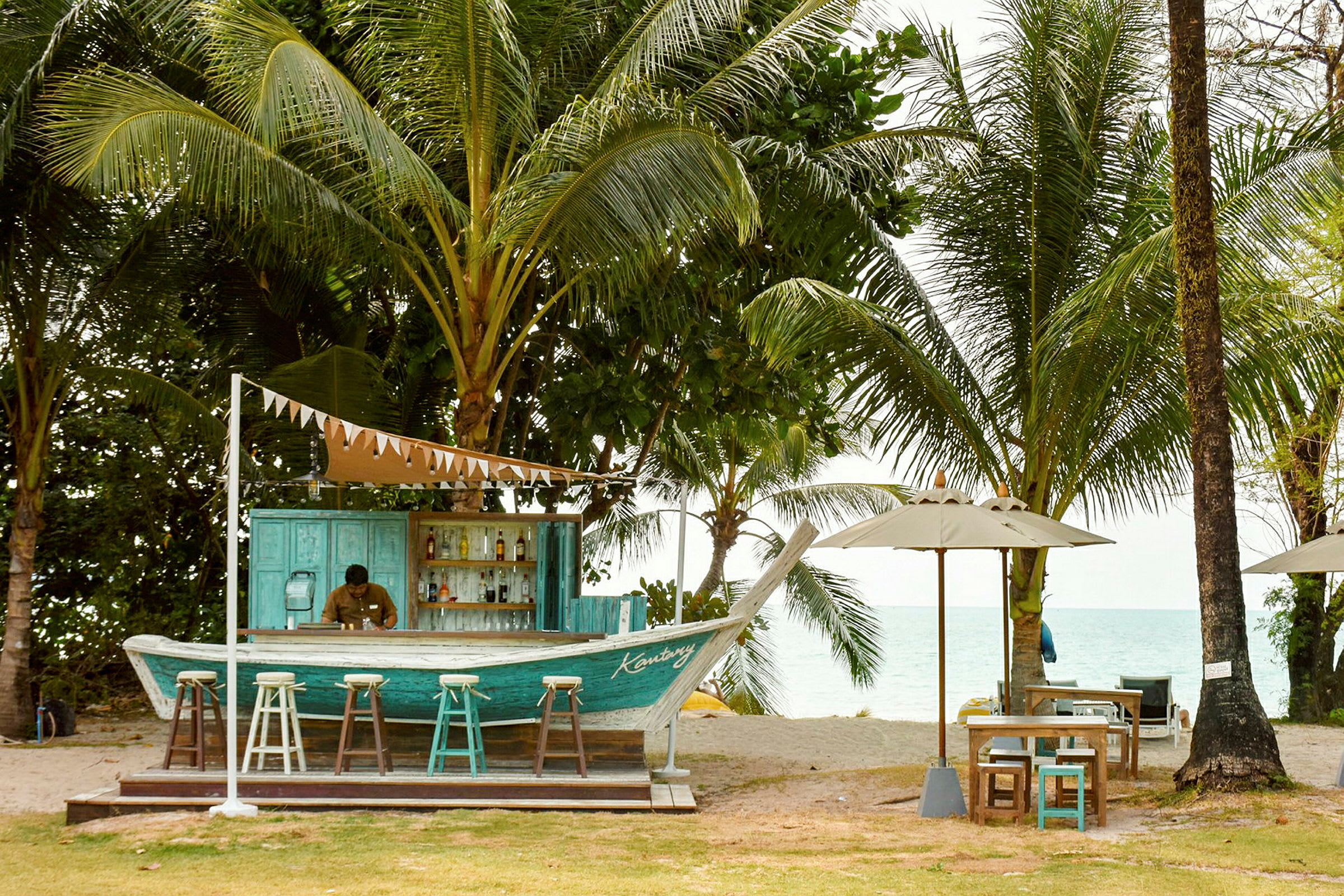 En strandbar byggd i form av en båt, omgiven av palmträd och med utsikt över havet. Det finns barstolar framför baren och bord med parasoll intill. (AI-genererad text)