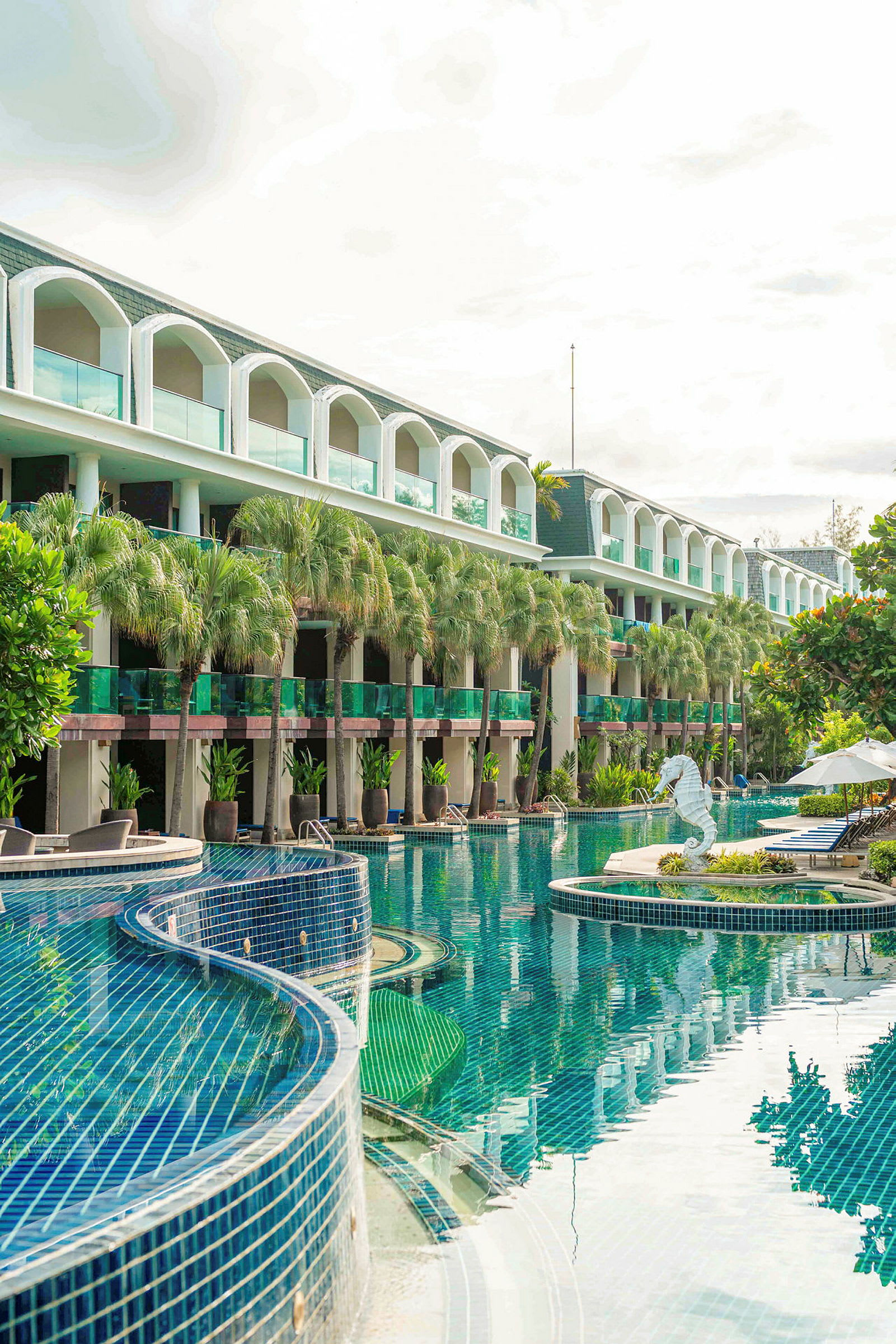 Bilden föreställer en pool utomhus omgiven av ett hotell med balkonger. Det finns gröna växter och en fontän i form av en sjöhäst. (AI-genererad text)