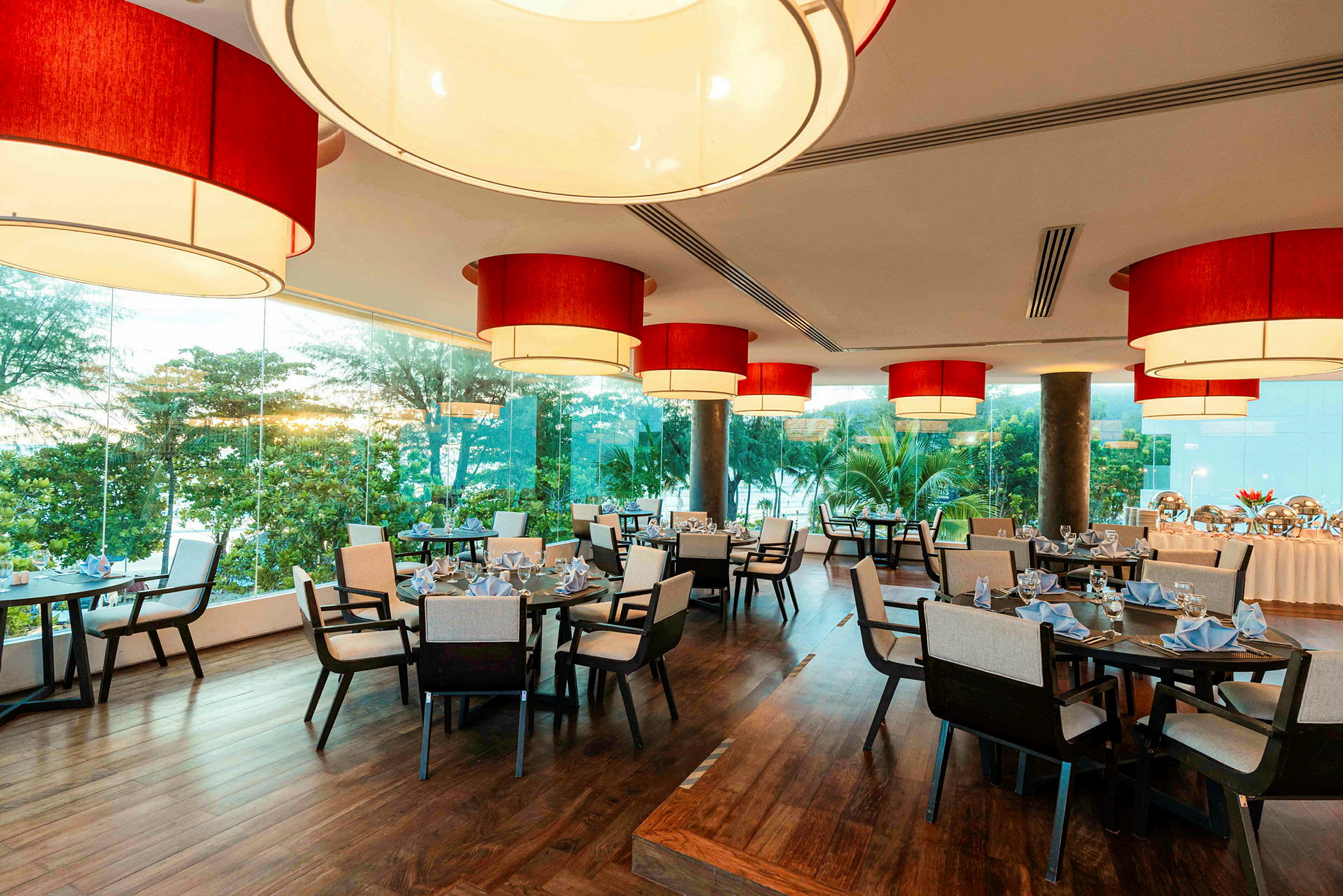 En restaurang med stora röda lampor i taket, fönster med utsikt över grönskande träd och dukade bord. (AI-genererad text)