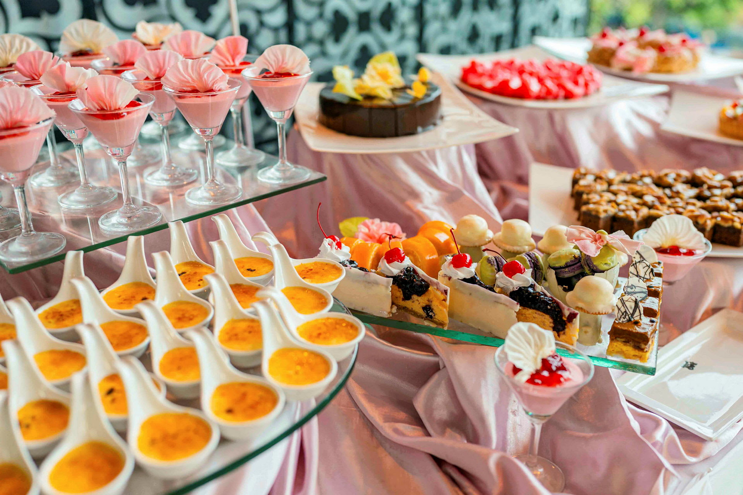 En buffé med olika desserter, inklusive glas med rosa mousse, crème brûlée, och dekorerade bakverk. (AI-genererad text)