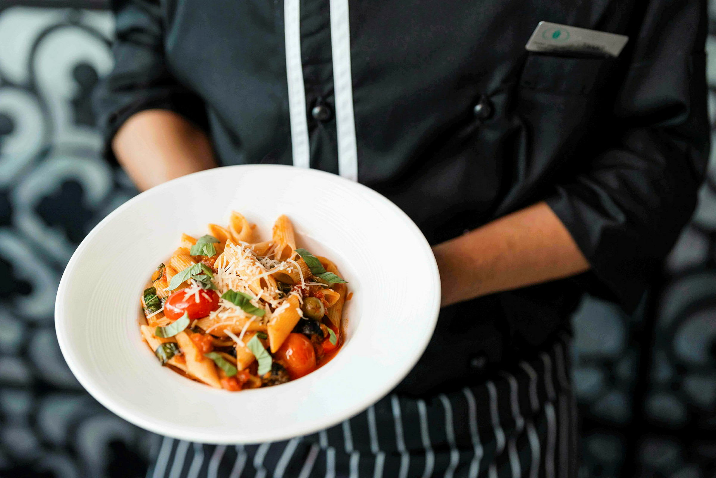 En person i kockkläder håller i en tallrik med en servering av pasta toppad med färska örter och ost. (AI-genererad text)