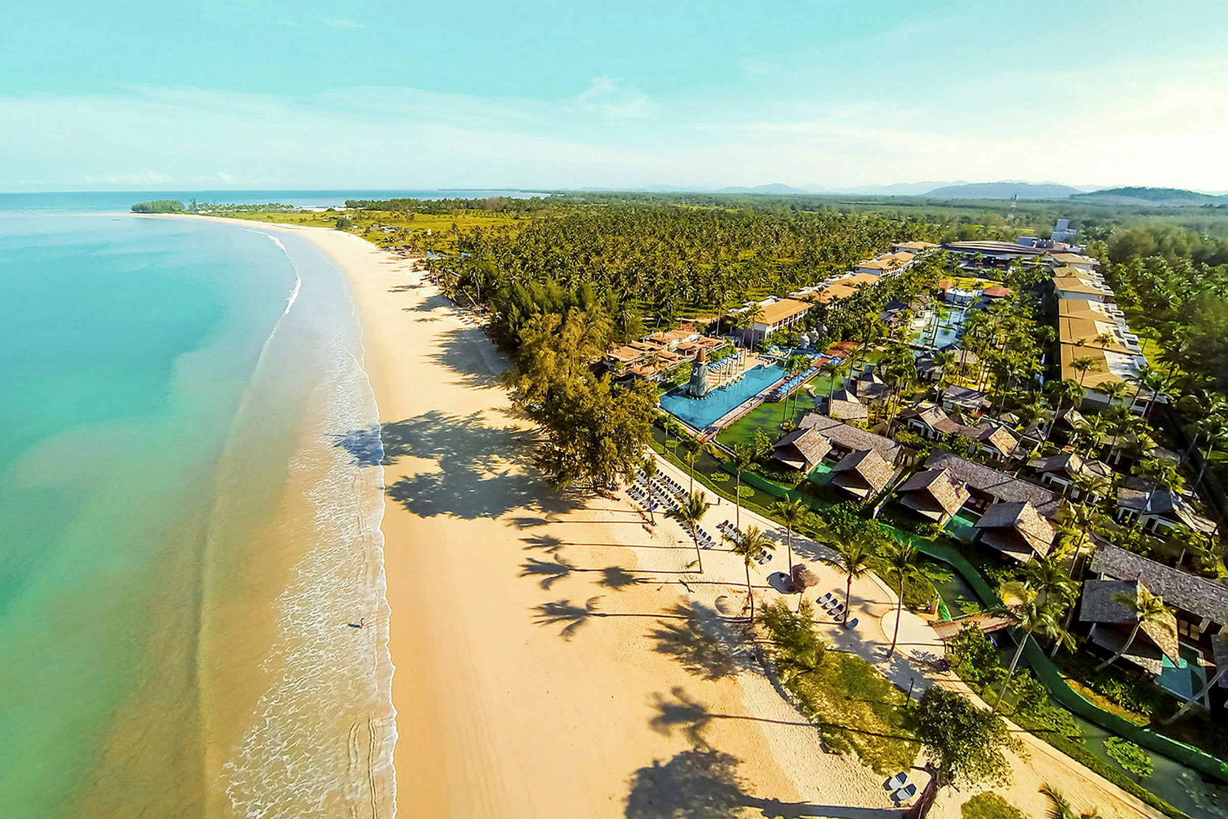 Flygfoto av en lång sandstrand med turkost vatten och en semesteranläggning med bungalower och pooler. (AI-genererad text)