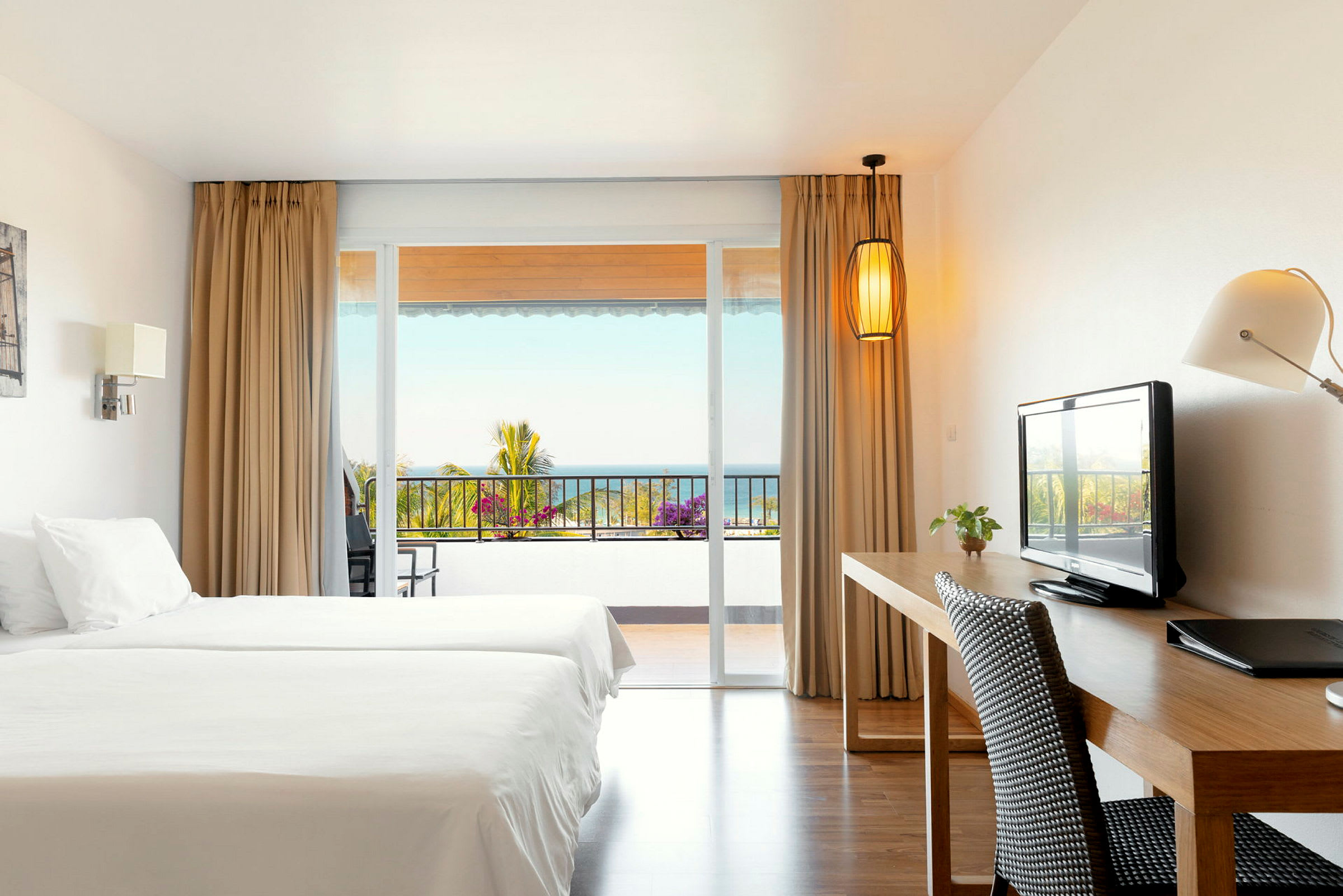 Ett hotellrum med två sängar, skrivbord, och utsikt mot en balkong med hav i bakgrunden. (AI-genererad text)